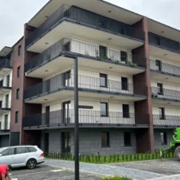 Nowoczesny budynek wielorodzinny z balkonami i elewacją z elementami cegły, widoczny z zewnątrz, z zaparkowanym samochodem i sprzętem budowlanym.