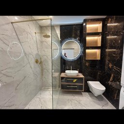 IZBUD - Nowoczesna łazienka z kabiną prysznicową typu walk-in, marmurowymi płytkami, okrągłym lustrem z podświetleniem LED i czarno-złotymi akcentami.