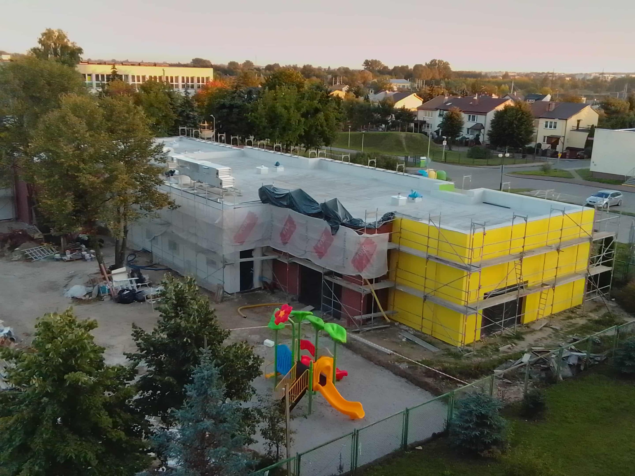 Budynek w trakcie ocieplania elewacji, widoczne rusztowania i plac zabaw w pobliżu. Część budynku ma żółtą elewację, a część jest w trakcie prac. Ujęcie z lotu ptaka.