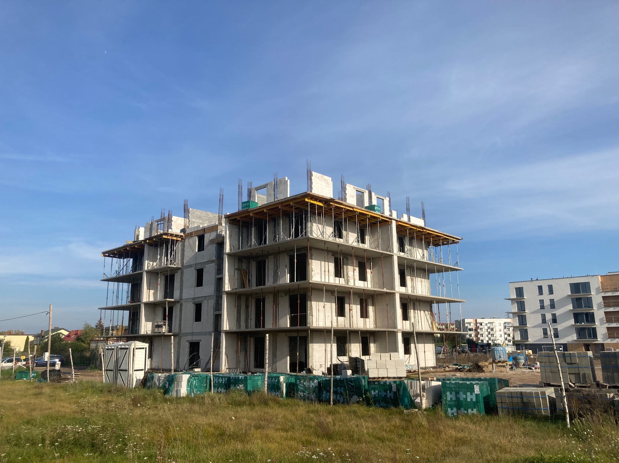 Wznoszony budynek wielorodzinny z białego bloczka, widoczne zbrojenia i szalunki stropów. Na pierwszym planie palety z materiałami budowlanymi i trawa.