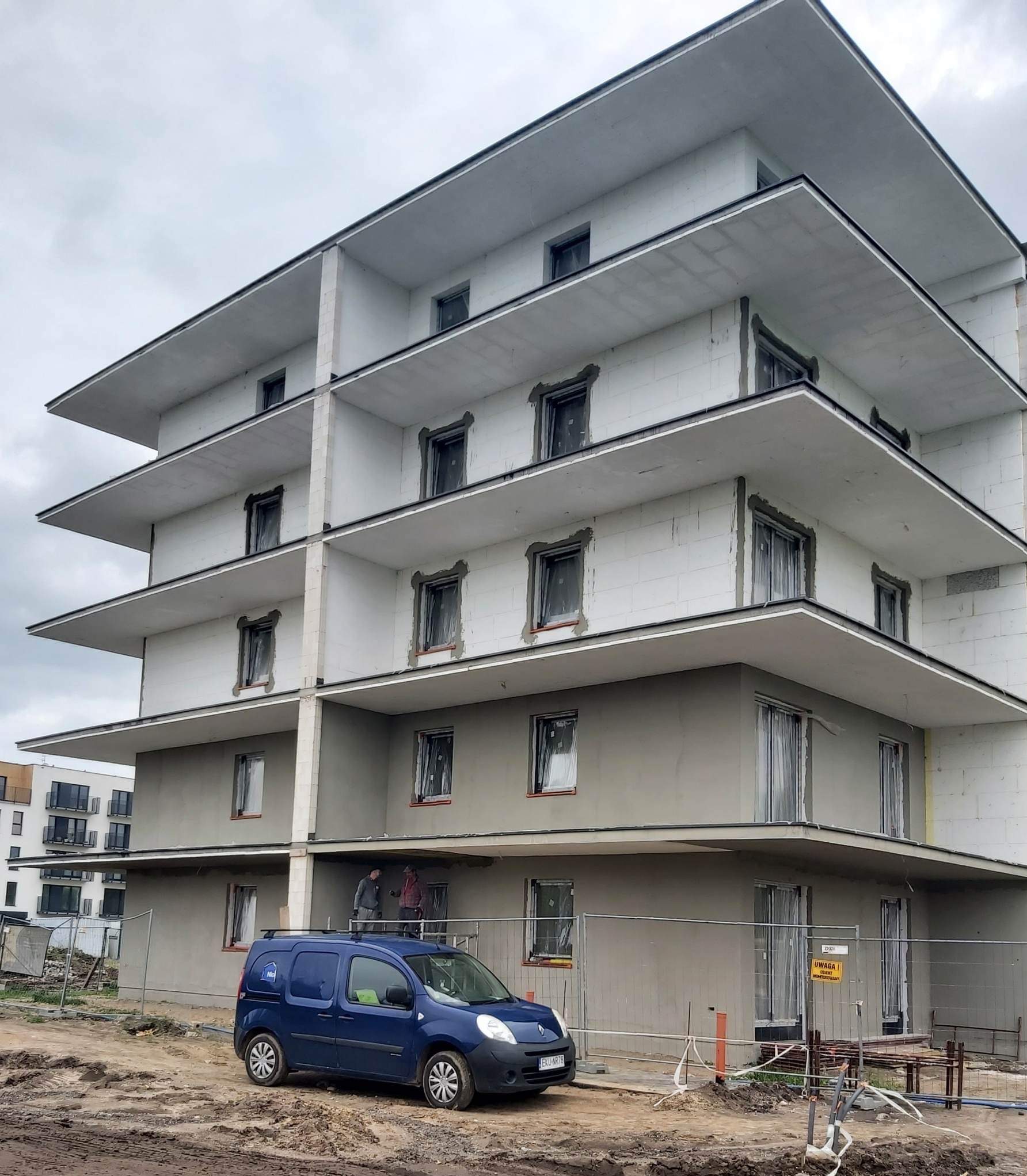Wielopiętrowy budynek w trakcie ocieplania elewacji, z widoczną warstwą izolacyjną i pracownikami na rusztowaniu. Na pierwszym planie niebieski samochód dostawczy.