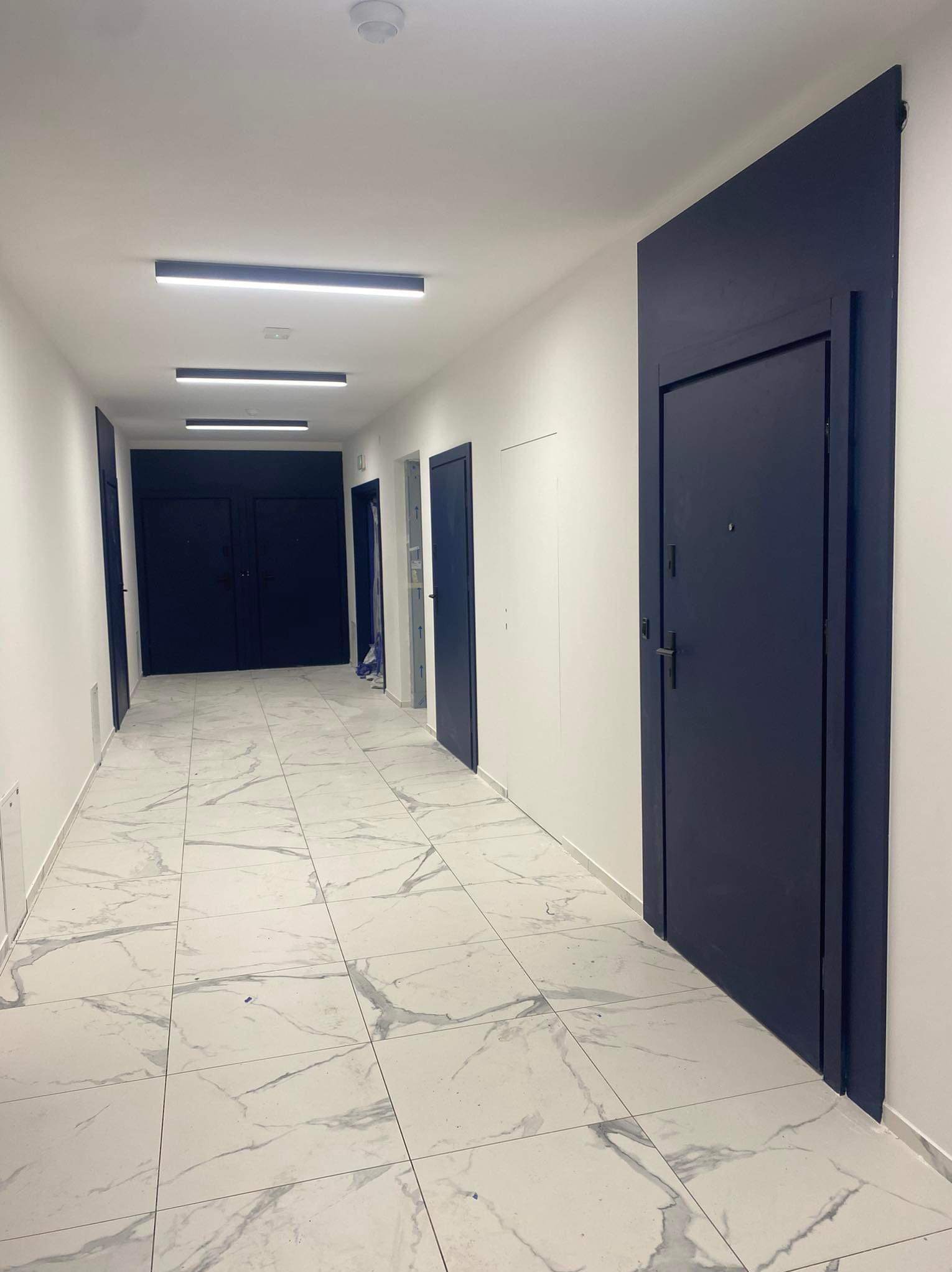 Wykończony korytarz z białymi ścianami, marmurową podłogą i ciemnymi drzwiami. Nowoczesne oświetlenie sufitowe. Perspektywa wnętrza ukazuje czystość i minimalizm.