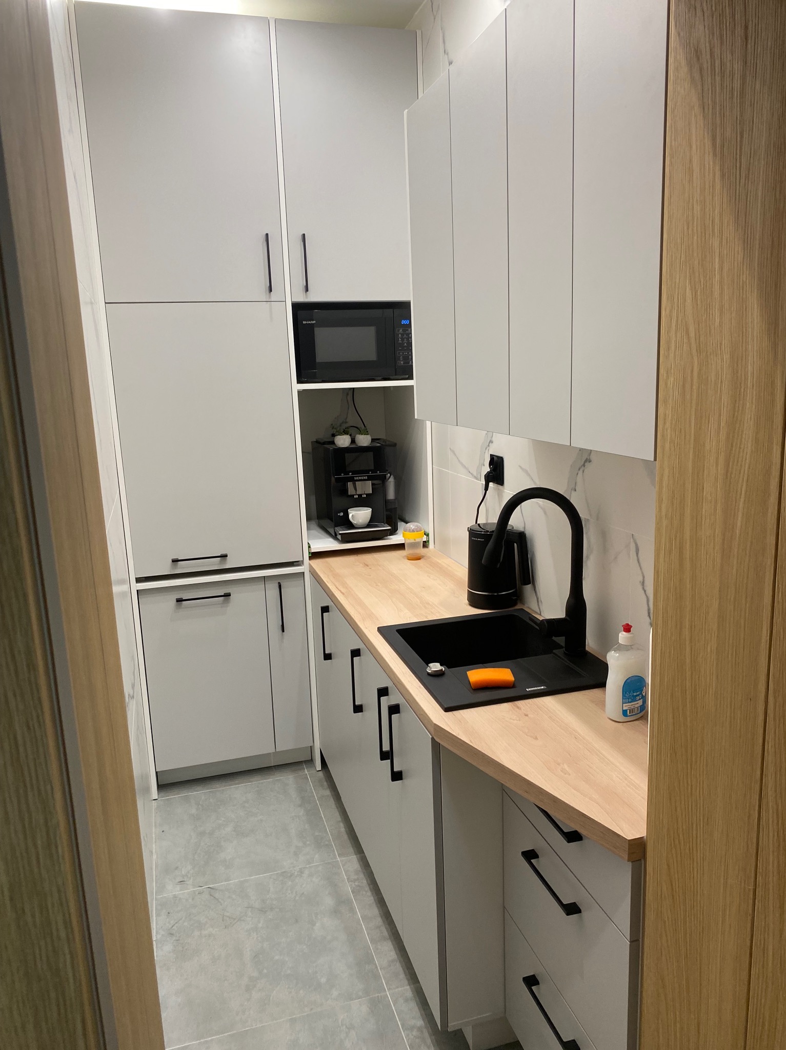 Nowoczesna kuchnia w odcieniach szarości i bieli z drewnianym blatem, czarną armaturą i sprzętem AGD. Minimalistyczny design, marmurowe płytki na ścianie.