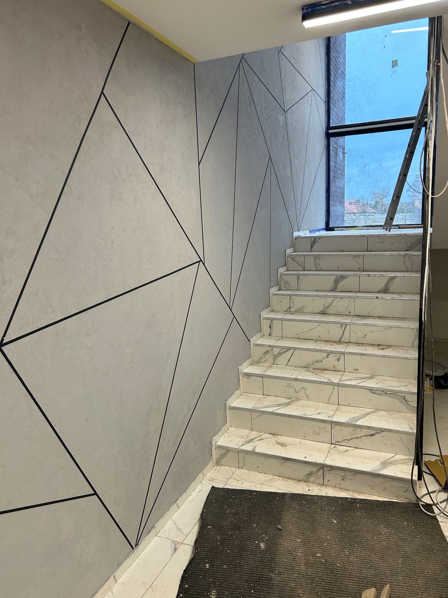 Schody wyłożone białymi płytkami imitującymi marmur prowadzą w górę, ściana z szarą dekoracyjną fakturą i czarnymi geometrycznymi liniami. Okno na końcu schodów.