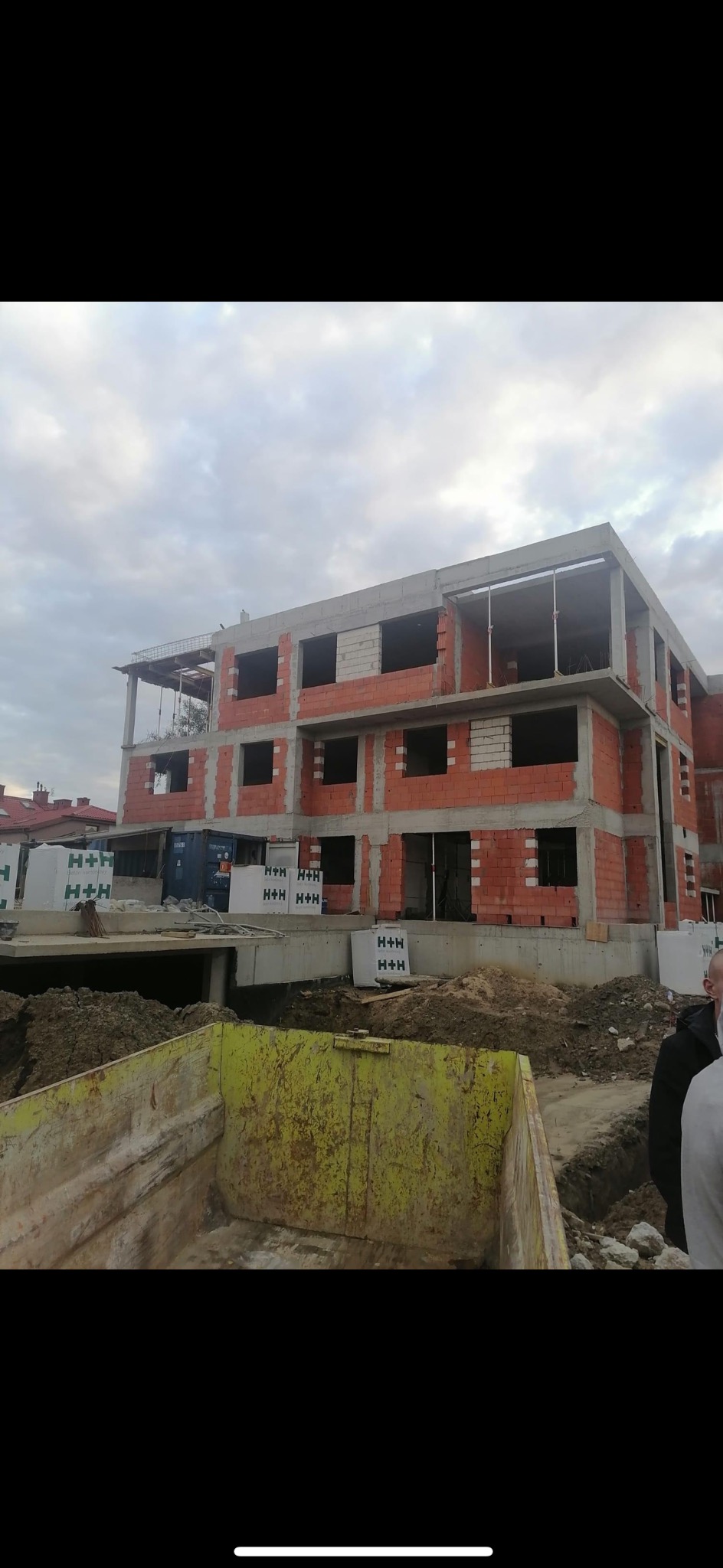 Wznoszenie trzypiętrowego budynku z czerwonej cegły H+H. Widoczne otwory okienne, szary betonowy strop i żółta skrzynia na gruz. Budowa w toku, pochmurne niebo.