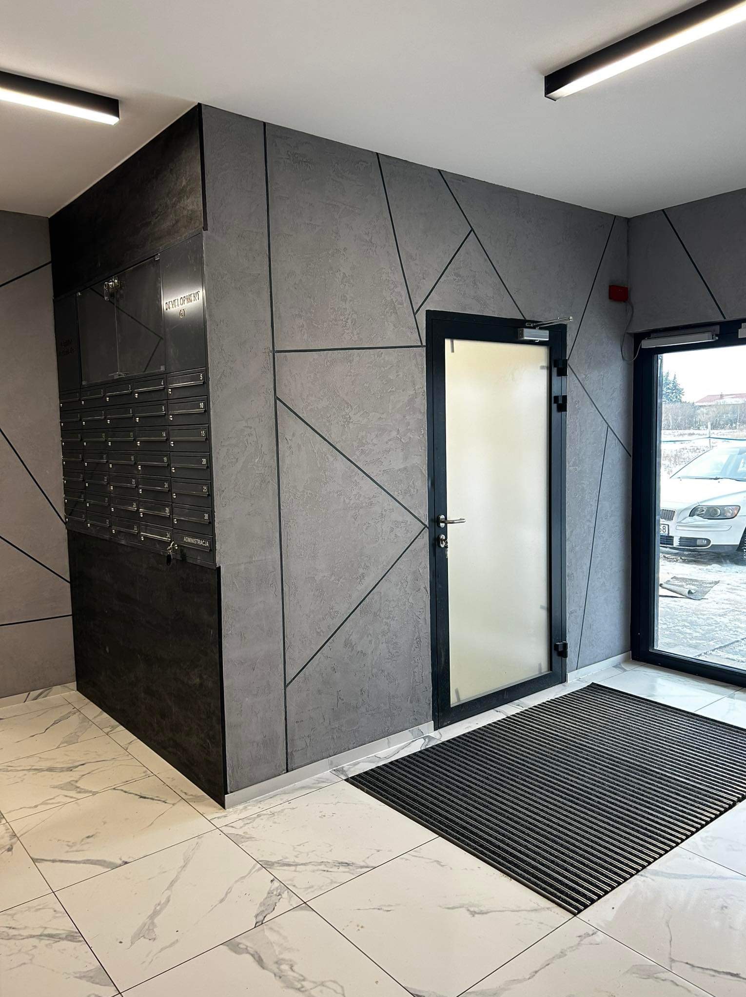 Nowoczesne wykończenie wnętrza klatki schodowej z szarą ścianą zdobioną geometrycznym wzorem, skrzynkami pocztowymi i marmurową podłogą. Widoczne drzwi wejściowe.