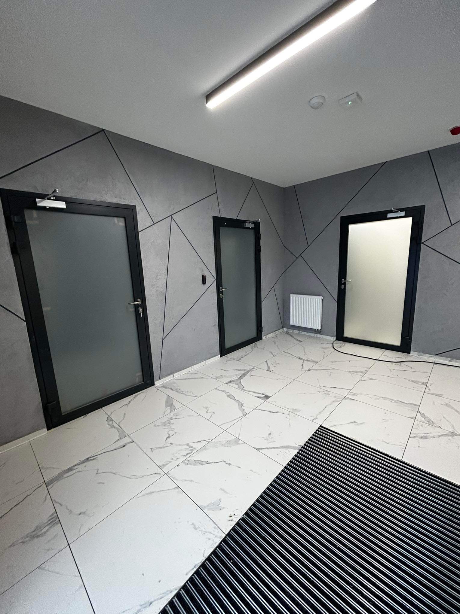 Wnętrze korytarza z trzema drzwiami z czarnymi ramami i matowym szkłem, podłoga z białych płytek imitujących marmur, ściany szare z geometrycznym wzorem.