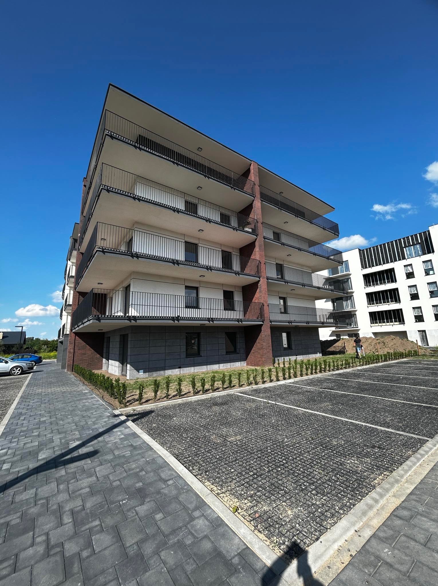 Nowoczesny, wielopiętrowy apartamentowiec z balkonami, wykończony cegłą i szarym kamieniem, na tle błękitnego nieba i częściowo wykończonego parkingu.