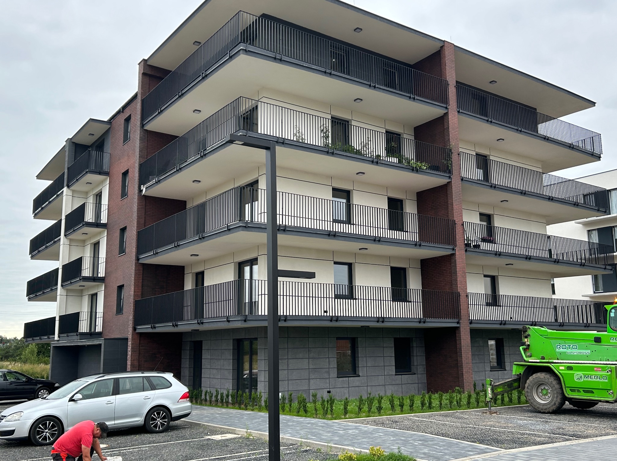 Nowoczesny budynek wielorodzinny z balkonami i elewacją z elementami cegły, widoczny z zewnątrz, z zaparkowanym samochodem i sprzętem budowlanym.