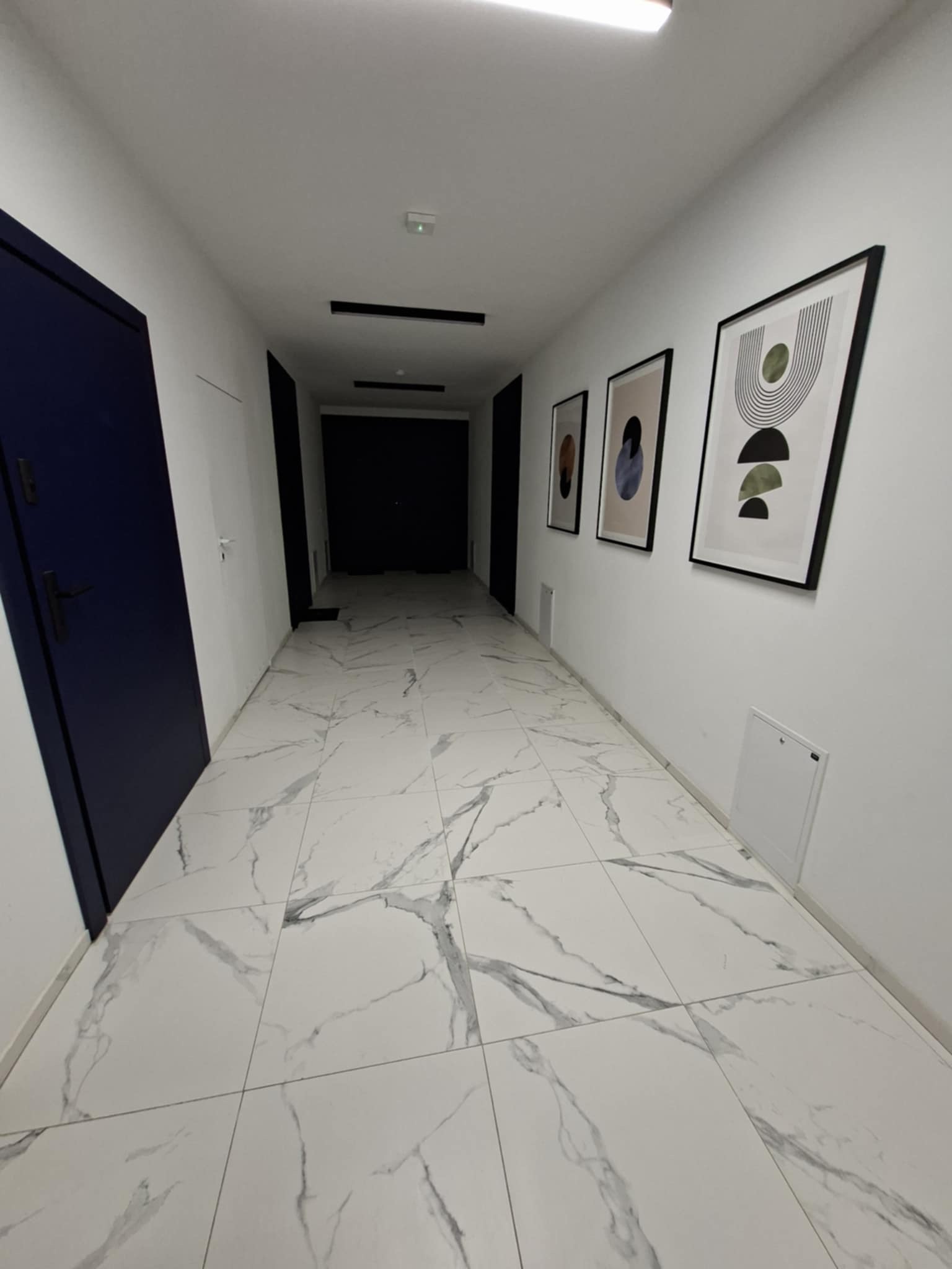 Długi korytarz z białymi płytkami imitującymi marmur, ciemnoniebieskie drzwi po lewej, minimalistyczne obrazy na ścianie po prawej. Nowoczesne wykończenie wnętrza.