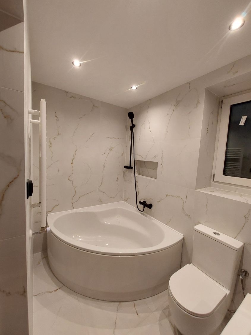 Wykończona łazienka z narożną wanną, czarną armaturą i białymi płytkami imitującymi marmur. Widoczne oświetlenie punktowe i grzejnik drabinkowy.