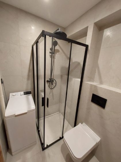 Nowoczesna łazienka z kabiną prysznicową typu walk-in z czarnymi profilami, białą toaletą podwieszaną i pralką. Jasne płytki na ścianach i podłodze.