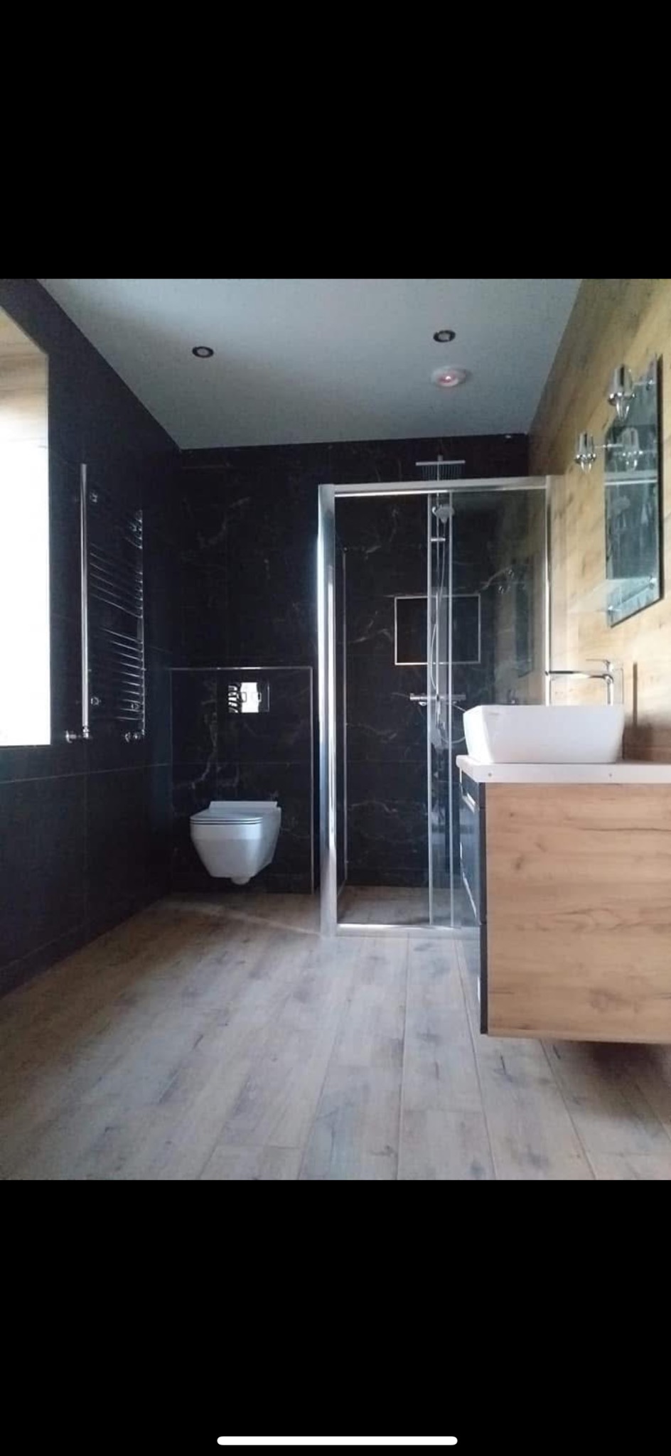 Nowoczesna łazienka po remoncie: czarne marmurowe ściany, drewnopodobna podłoga, kabina prysznicowa ze szkła i umywalka na drewnianej szafce.