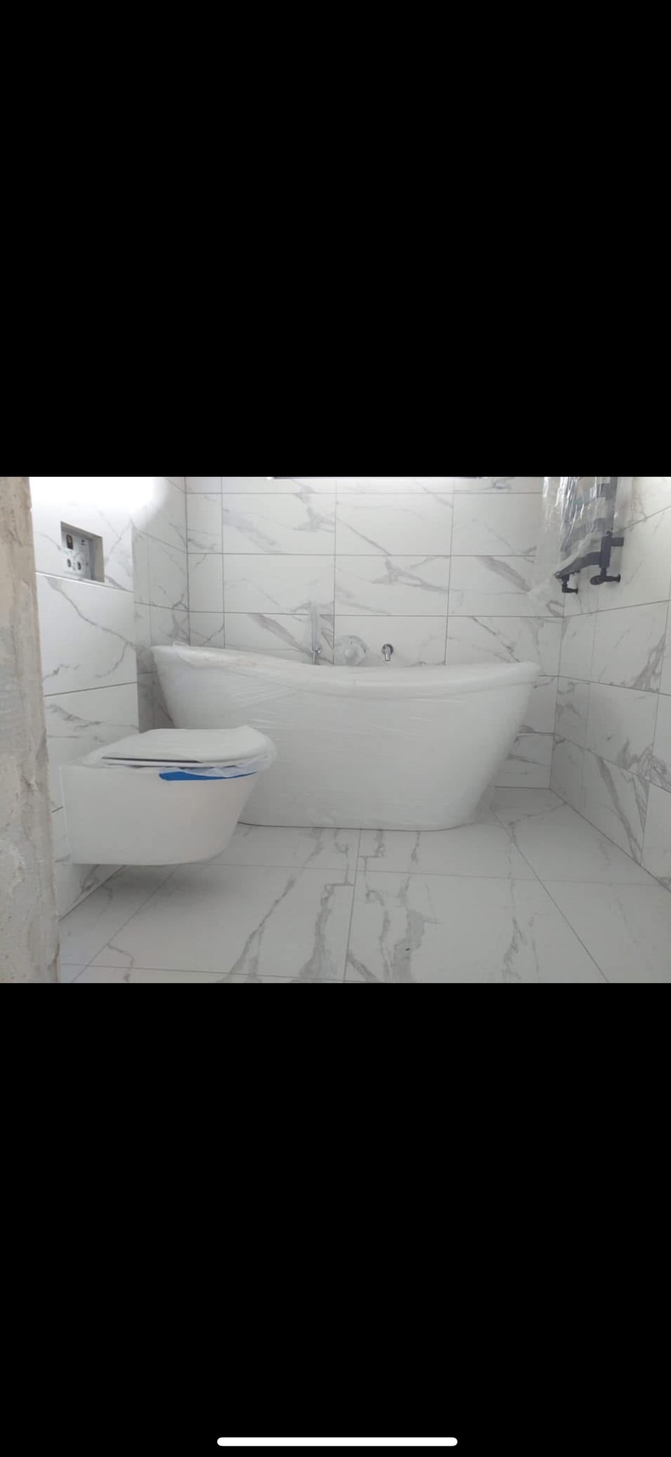 Biała, wolnostojąca wanna i podwieszana toaleta w łazience wyłożonej marmurowymi płytkami. Armatura i ceramika zabezpieczona folią ochronną.