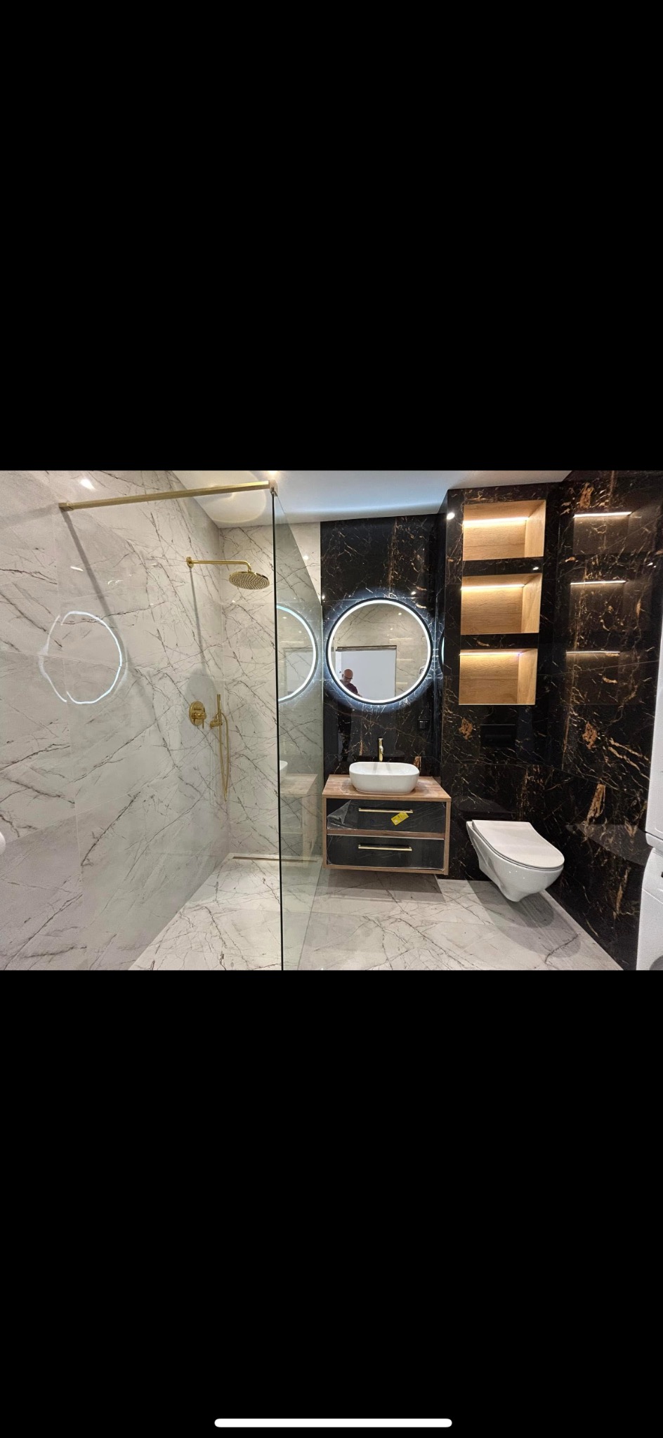 Nowoczesna łazienka z kabiną prysznicową typu walk-in, marmurowymi płytkami, okrągłym lustrem z podświetleniem LED i czarno-złotymi akcentami.