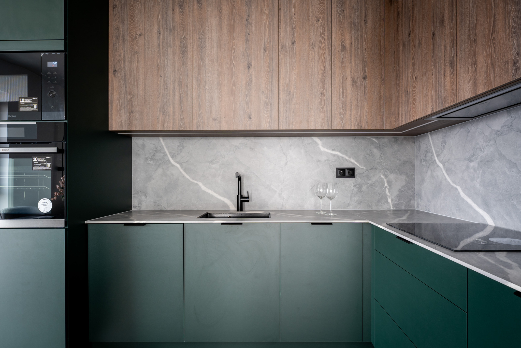 Nowoczesna kuchnia z zielonymi frontami, drewnianymi szafkami górnymi i marmurowym blatem. Minimalistyczny design, czarna armatura i sprzęt AGD. Dwa kieliszki do wina.
