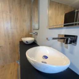 Nowoczesna łazienka z umywalkami Cersanit na czarnym blacie, drewnopodobne płytki ścienne, lustra z oświetleniem LED i chromowana armatura.