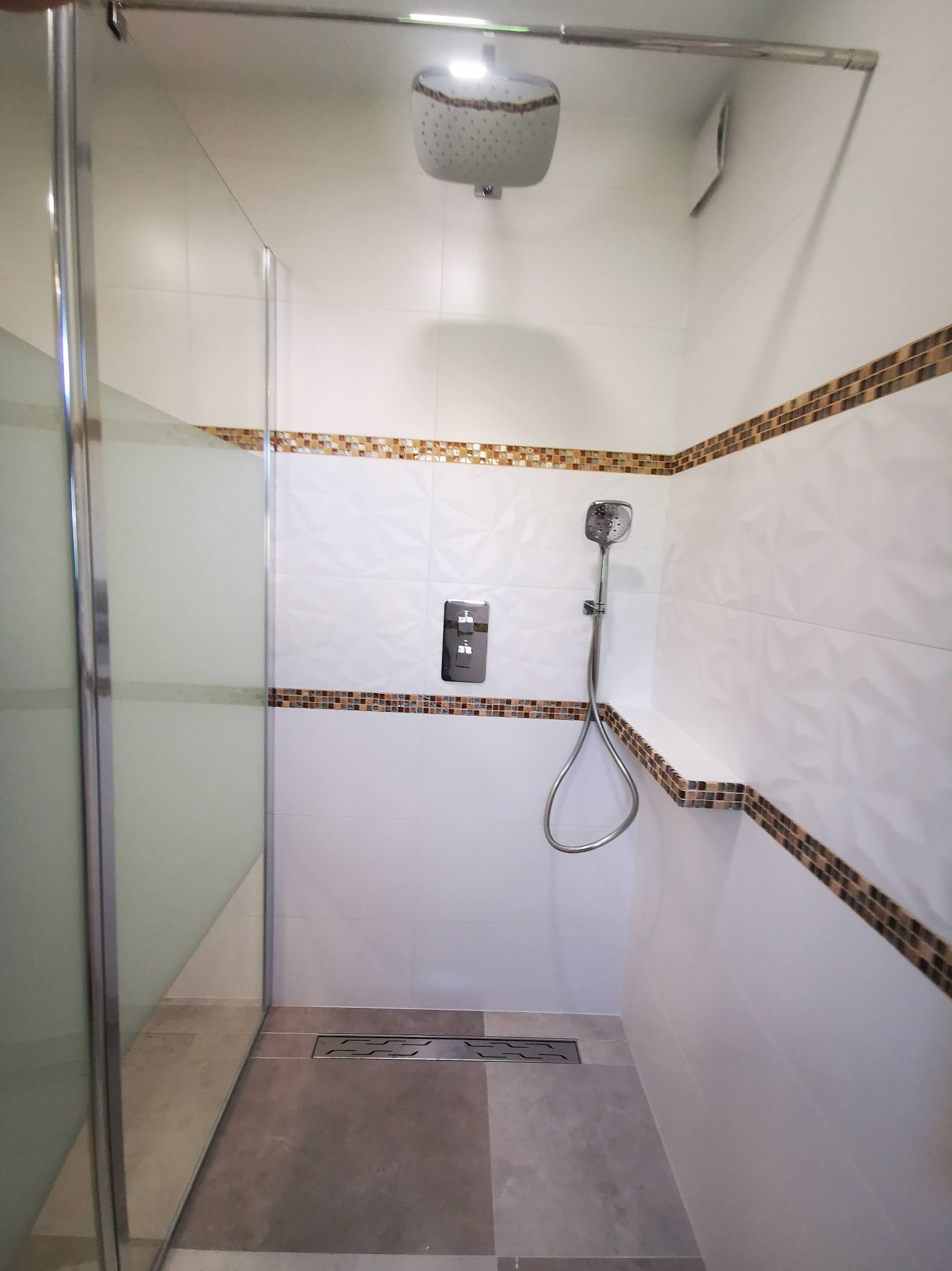 Kabina prysznicowa z deszczownicą i odpływem liniowym, wykończona białymi płytkami z dekoracyjnym pasem mozaiki. Nowoczesna armatura i szklane drzwi.