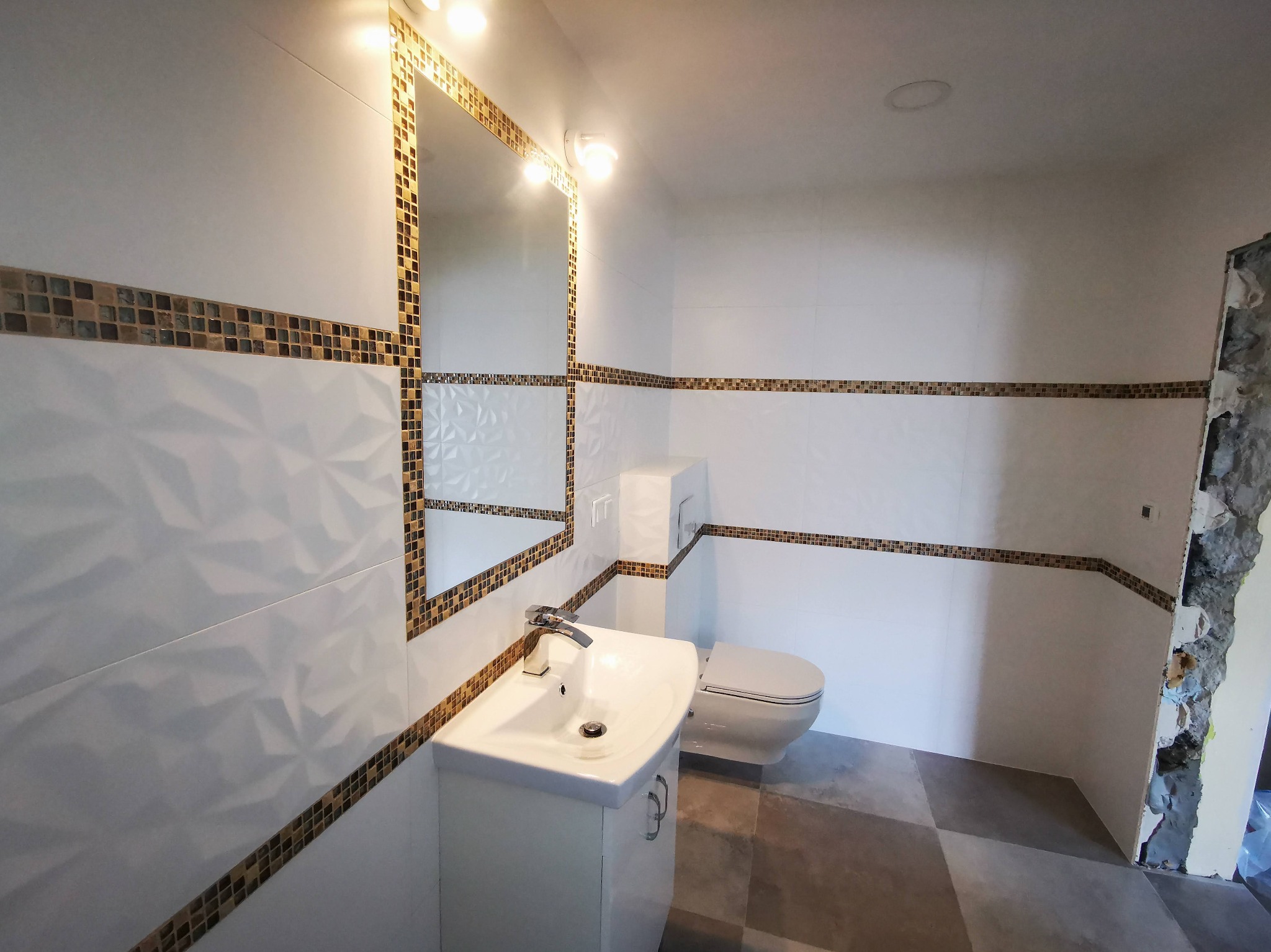 Wykończona łazienka z białą armaturą, mozaiką i dekoracyjnymi płytkami 3D. Częściowo wykończone ściany z odsłoniętym murem. Umywalka z szafką, lustro z oświetleniem.