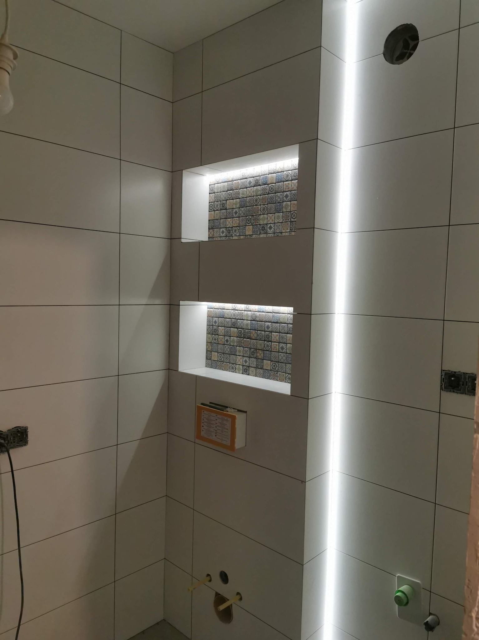 Białe, matowe płytki w łazience z wnękami podświetlonymi LED i dekoracyjnymi płytkami. Widoczne niedokończone instalacje i otwory na oświetlenie.