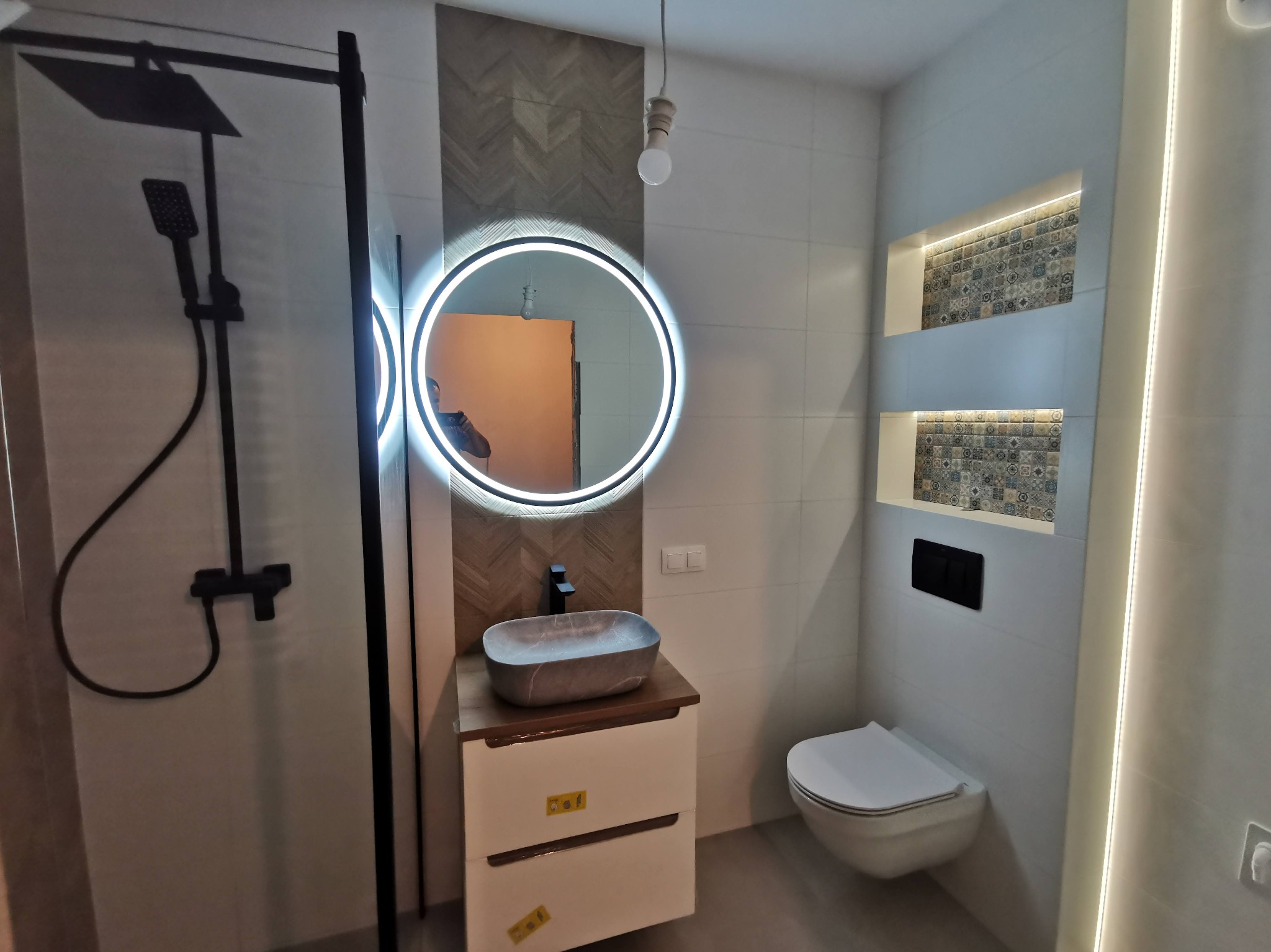 Nowoczesna łazienka z kabiną prysznicową typu walk-in, okrągłym lustrem z podświetleniem LED i umywalką nablatową. Na ścianach jasne płytki i dekoracyjne wnęki.