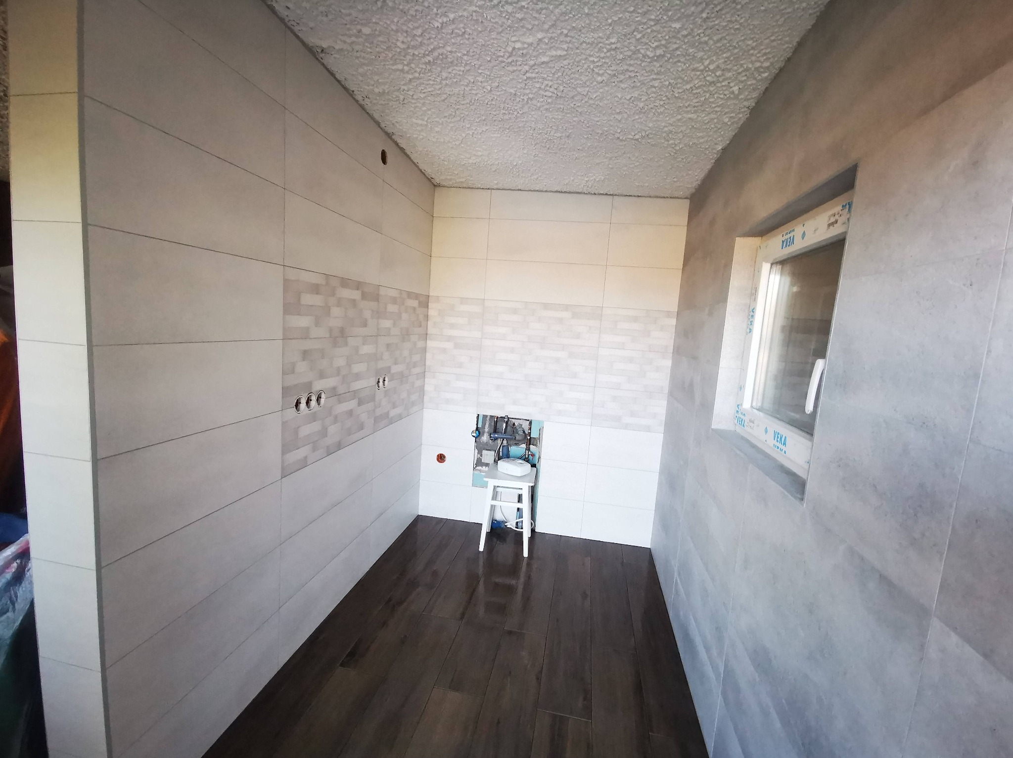 Łazienka po remoncie: ciemne panele podłogowe imitujące drewno, ściany wyłożone jasnymi, prostokątnymi płytkami z dekoracyjnym pasem mozaiki i oknem.