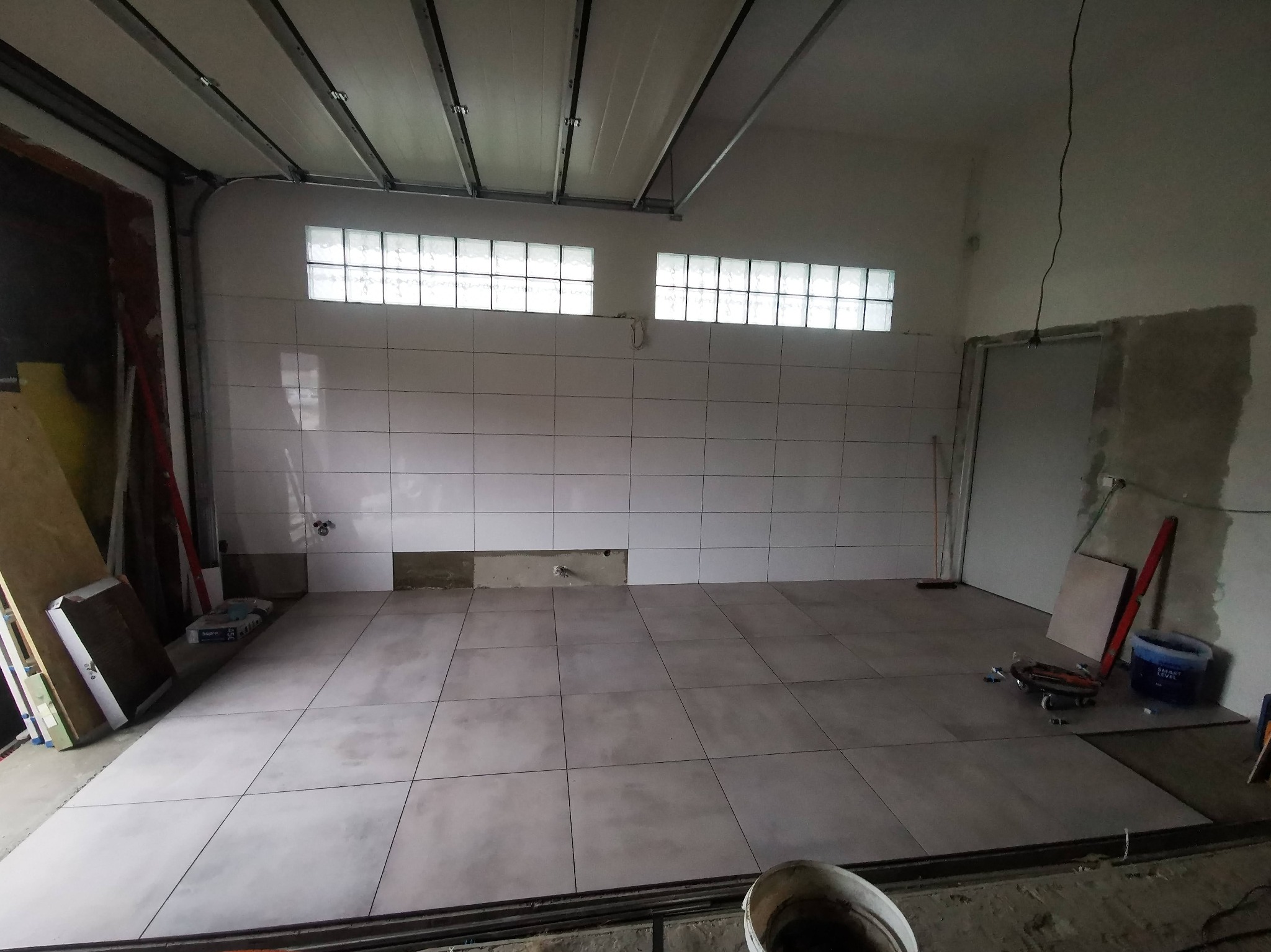 Układanie płytek podłogowych w garażu w Kędzierzynie-Koźlu. Widoczne narzędzia glazurnicze i fragment ściany z szklanymi luksferami.