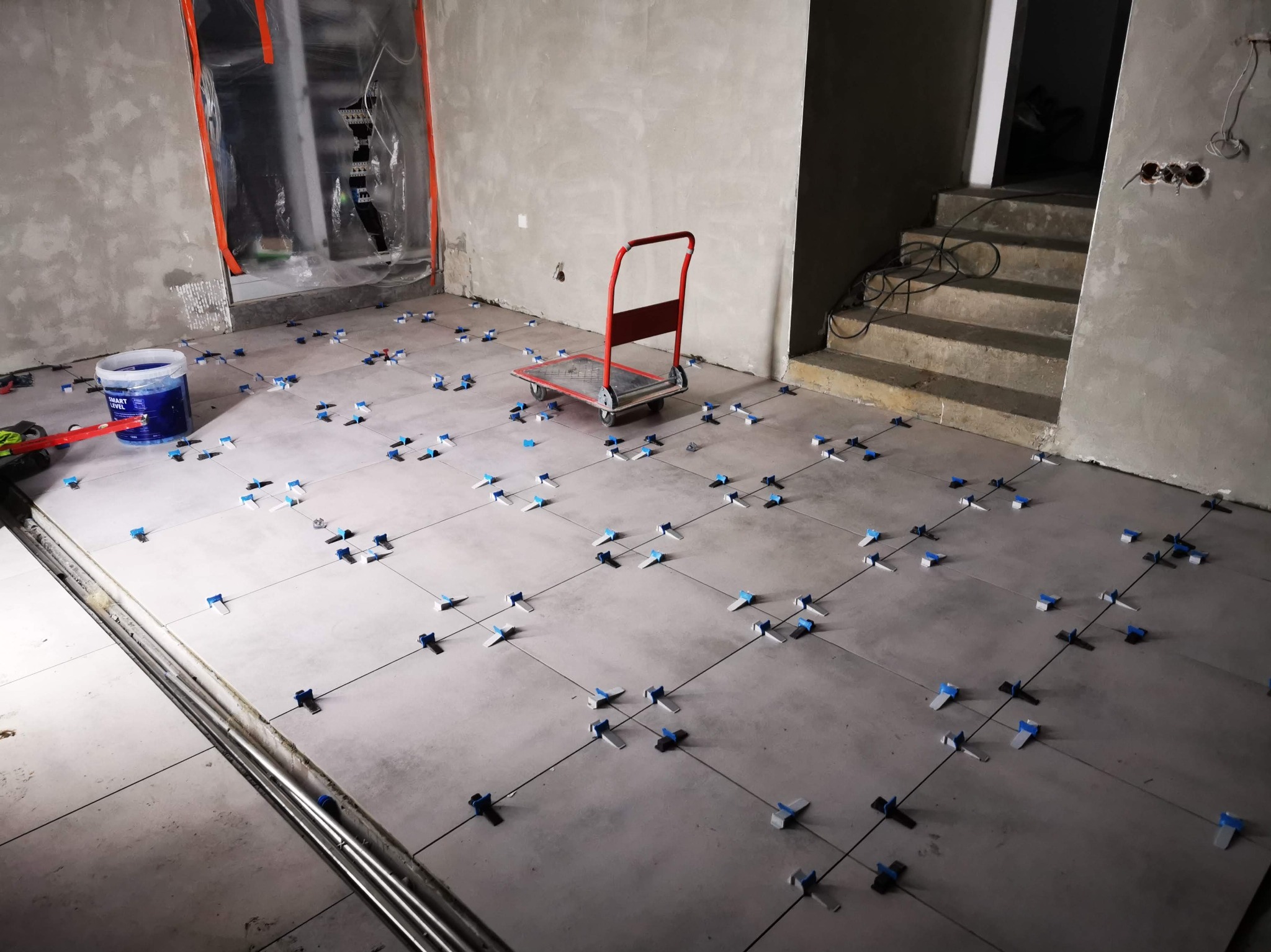 Szare płytki podłogowe w trakcie układania z użyciem niebieskich krzyżyków dystansowych. W tle wózek transportowy, schody i ściany w stanie surowym.