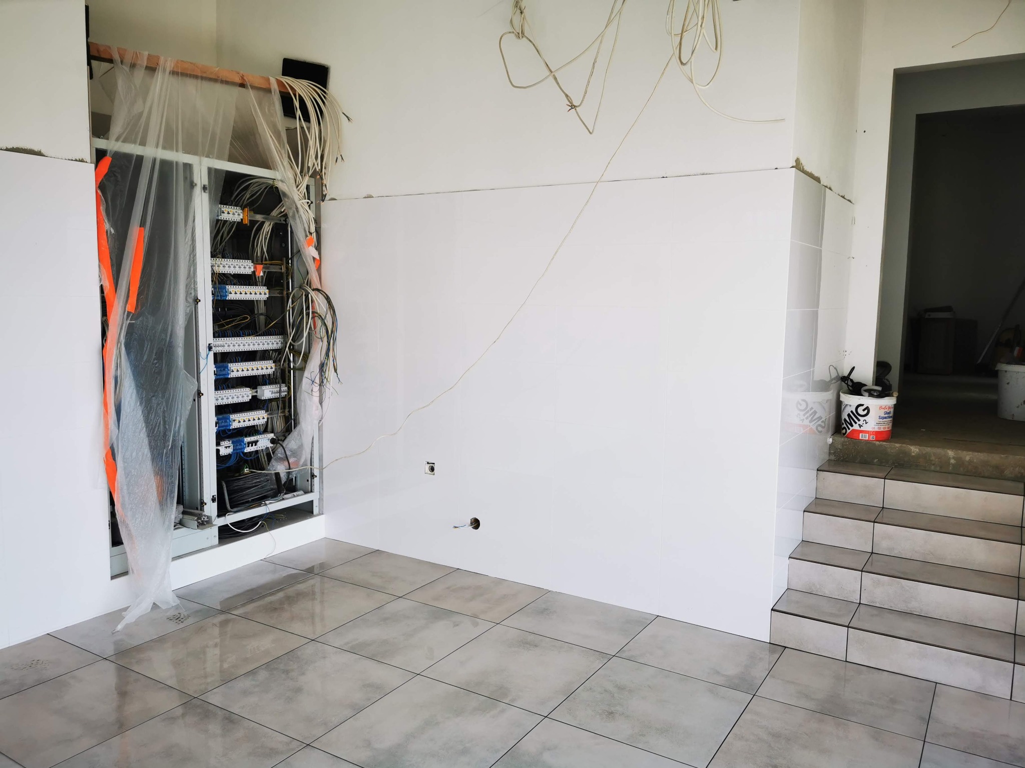 Wnętrze w trakcie prac: białe płytki na ścianach, szare płytki podłogowe, otwarta szafa elektryczna z okablowaniem. Widoczne schody z wykończeniem płytkami.
