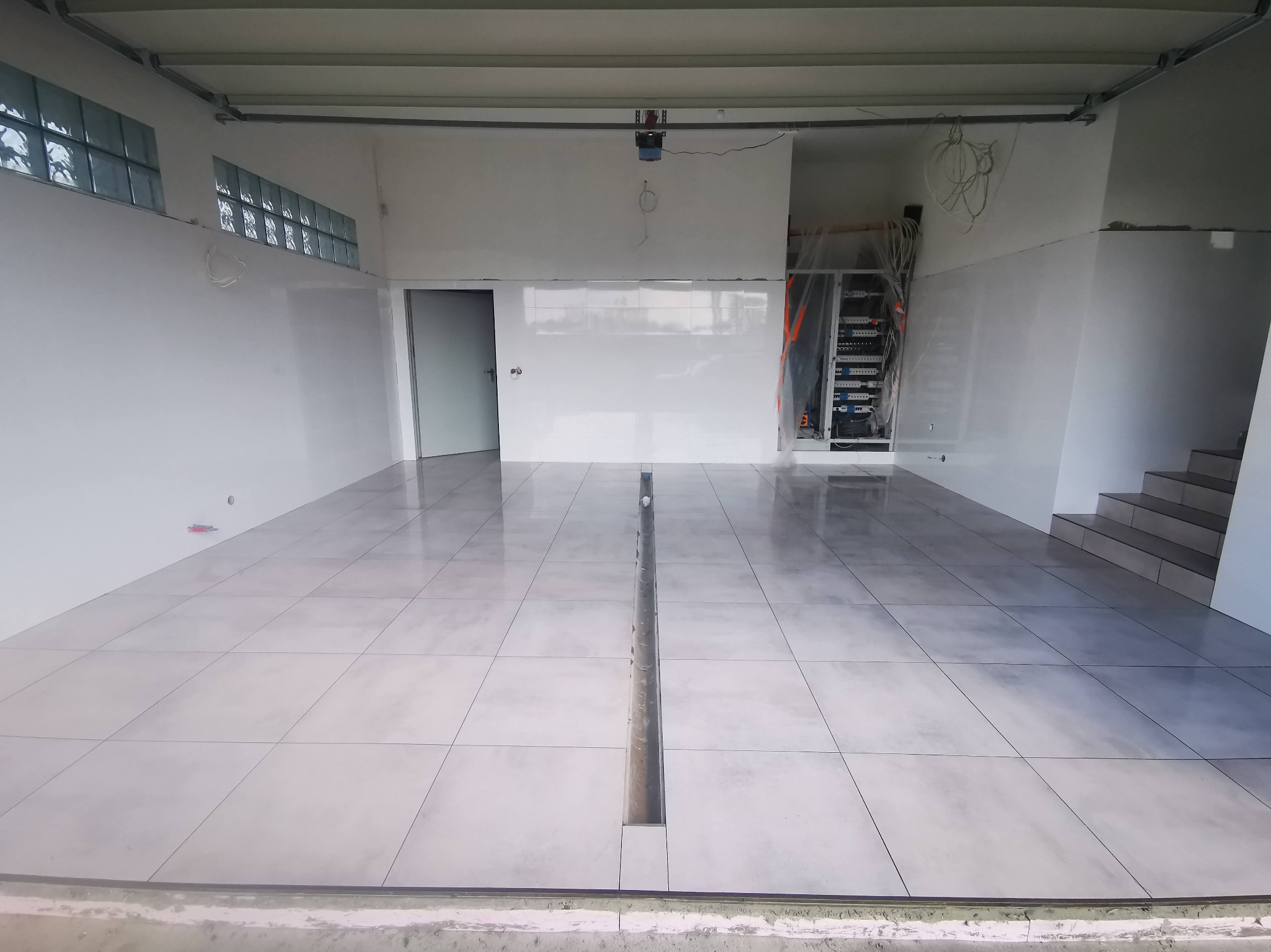 Wykończona posadzka z szarych płytek w jasnym pomieszczeniu z odpływem liniowym. Widoczne szklane luksfery w oknach i schody. Świeżo położone płytki podłogowe.