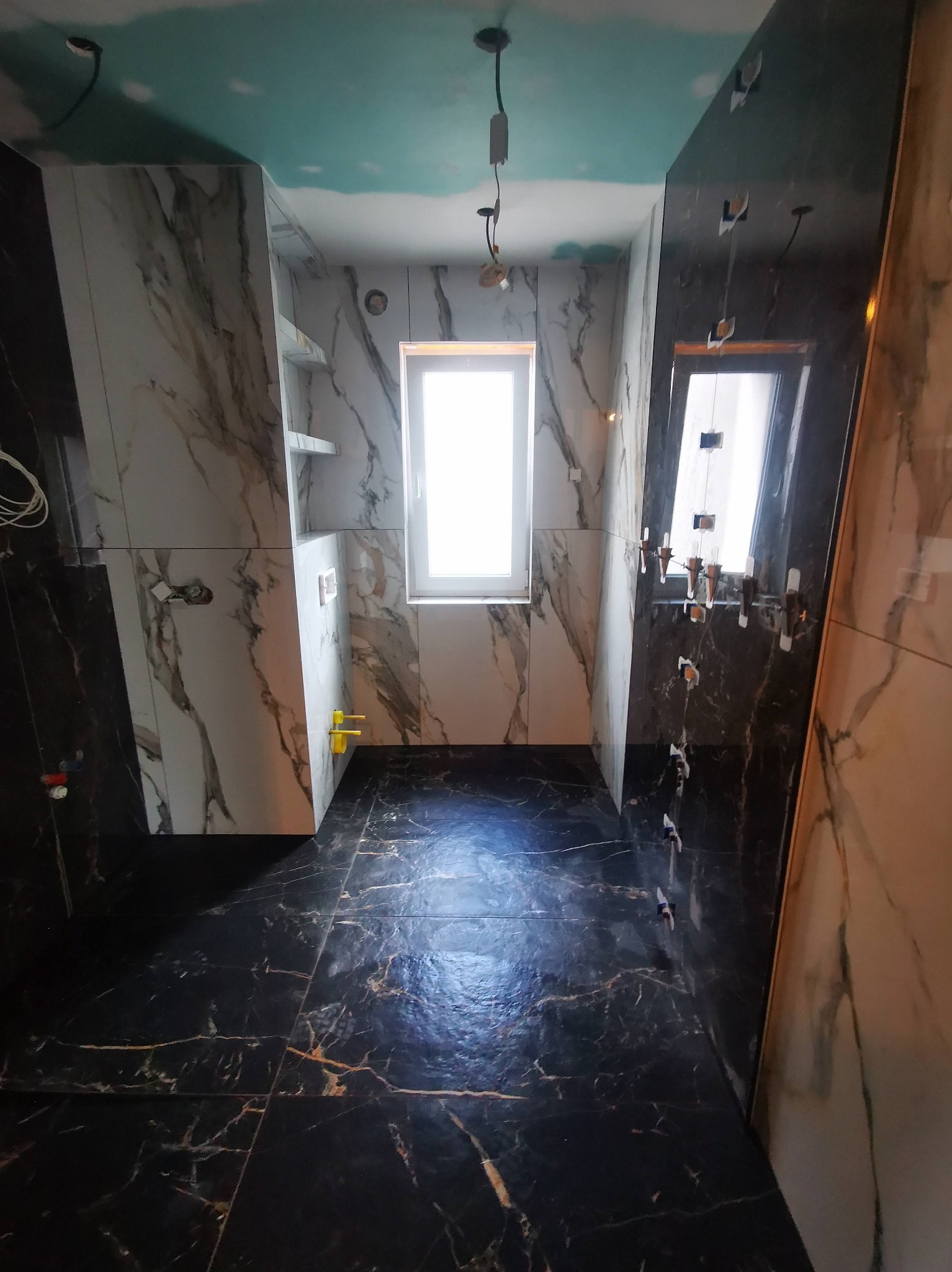 Wnętrze łazienki w trakcie wykańczania: marmurowe płytki na ścianach i podłodze, widoczne uchwyty do montażu lustra, niebieski sufit, okno w tle.
