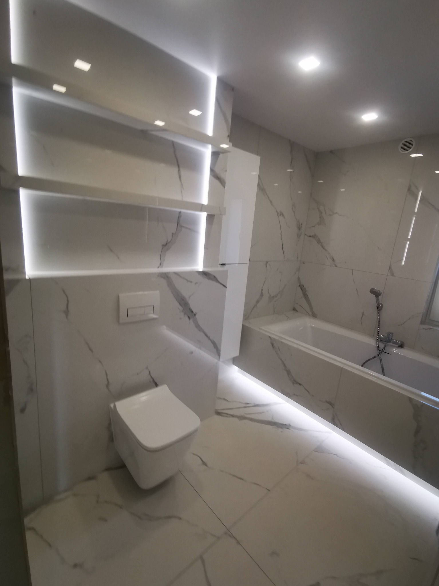 Nowoczesna łazienka z białymi płytkami imitującymi marmur, podświetlane półki i podłoga, wisząca toaleta. Minimalistyczny design z akcentem światła.
