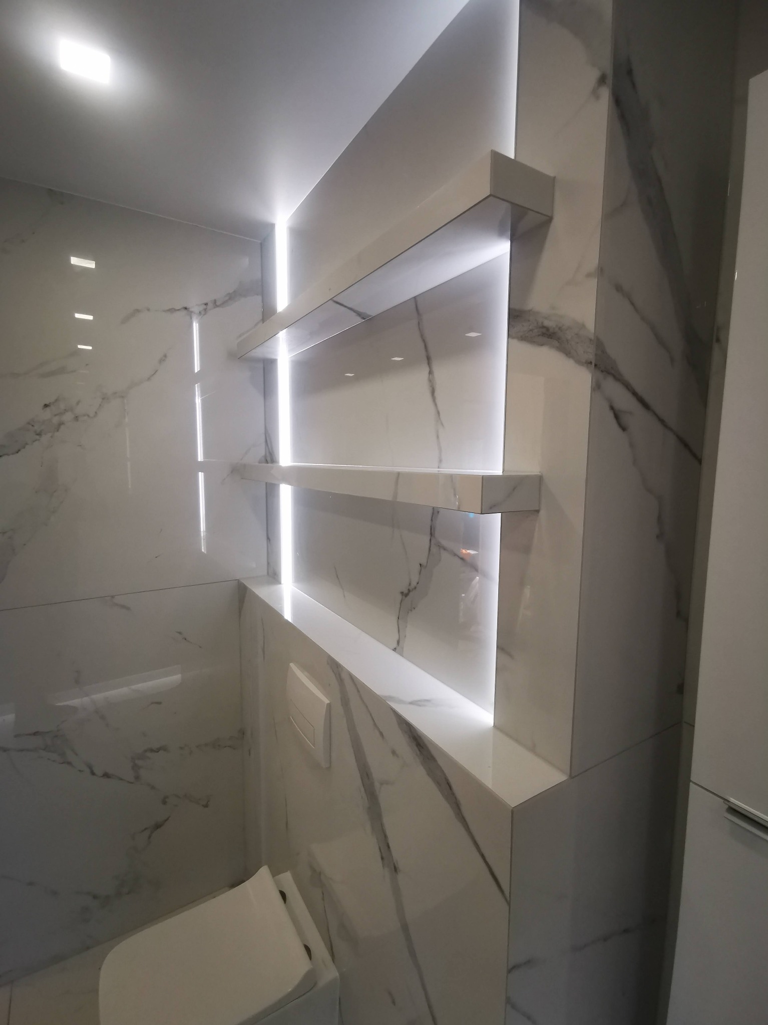 Elegancka łazienka w bieli: marmurowe płytki, podświetlane półki i wisząca toaleta tworzą nowoczesny, minimalistyczny design. Subtelne oświetlenie dodaje luksusowego charakteru.