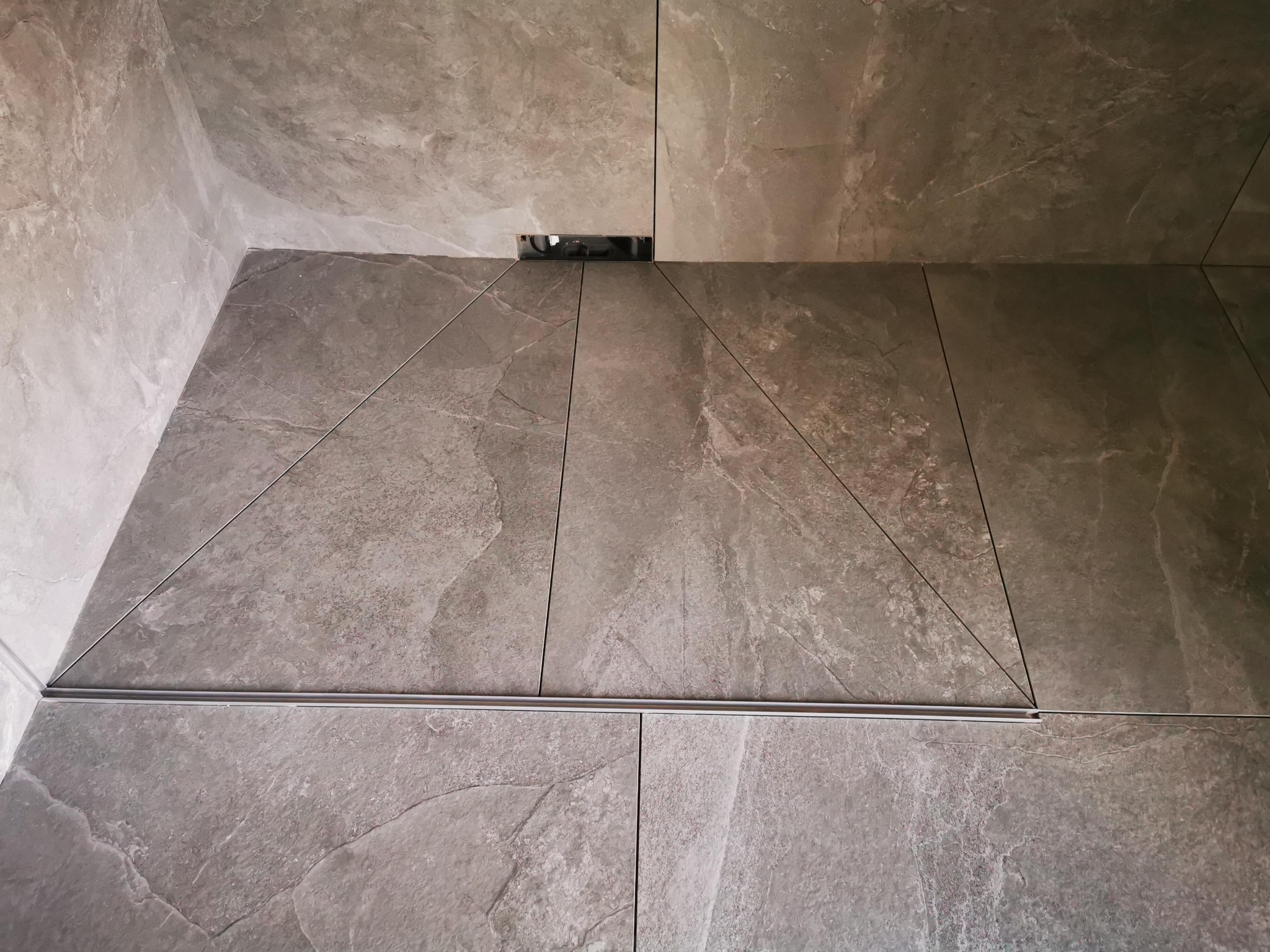 Szare płytki imitujące kamień, ułożone na podłodze i ścianie, z widocznym odpływem liniowym i listwą dekoracyjną w rogu pomieszczenia.