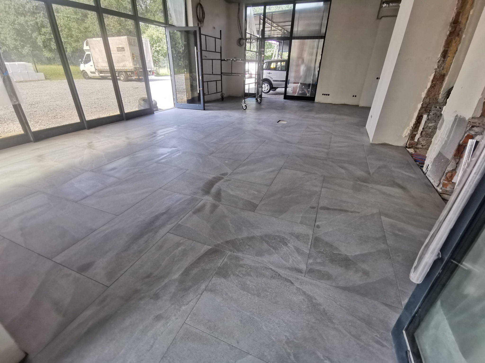 Szara, matowa podłoga z płytek w dużym pomieszczeniu z otwartymi drzwiami i surowymi ścianami. Widoczne elementy rusztowania i pojazd na zewnątrz.