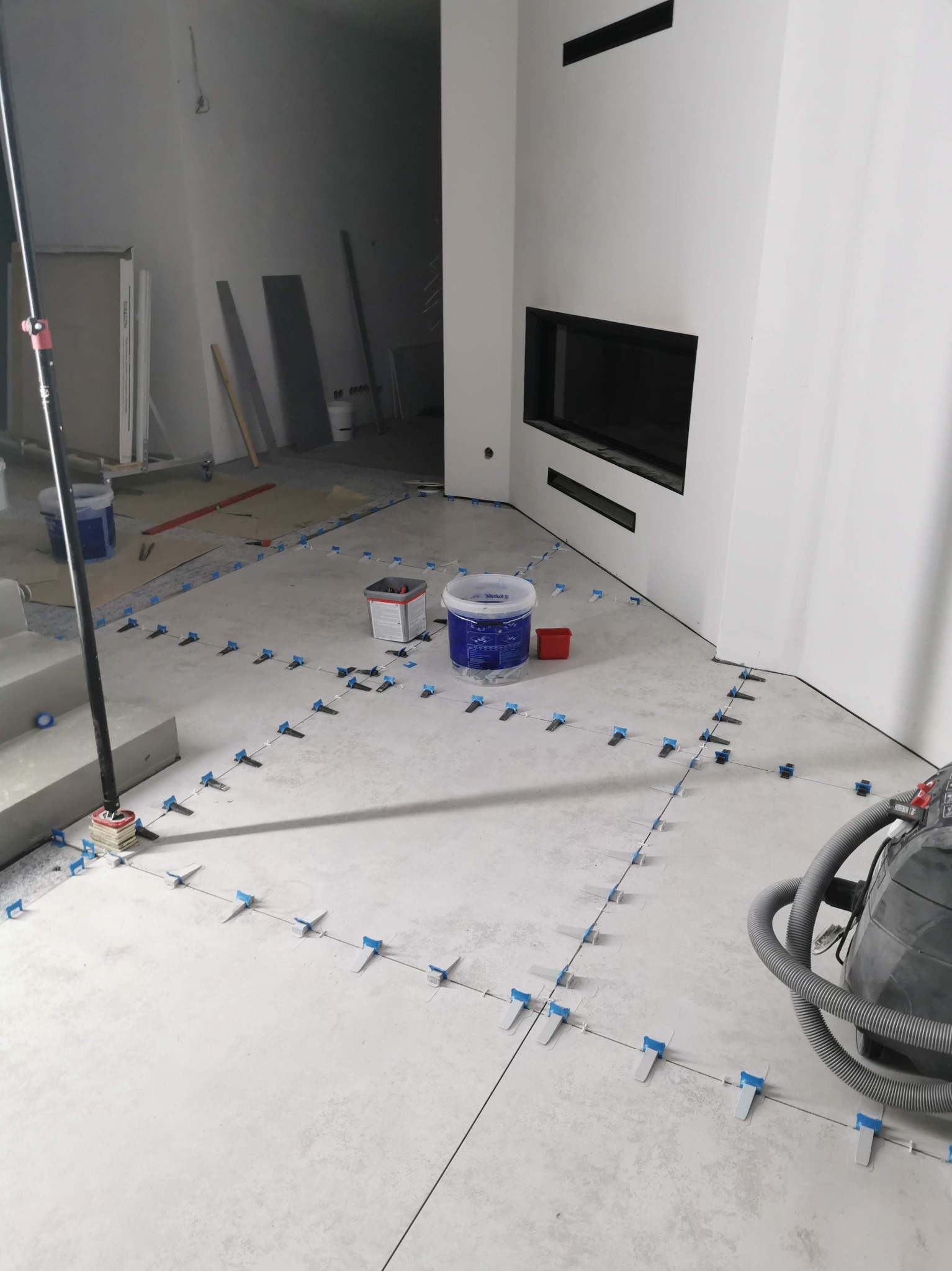 Układanie jasnych płytek podłogowych z użyciem systemu poziomowania. W tle widoczny kominek i przygotowane materiały budowlane.