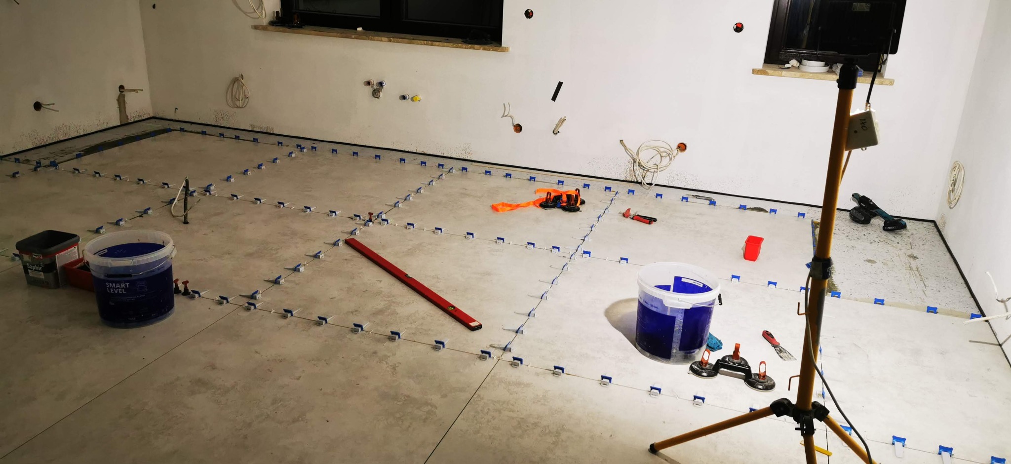 Etap prac: Wyrównywanie płytek podłogowych w jasnym pomieszczeniu za pomocą systemu poziomowania. Widoczne narzędzia, poziomica, wiadra i lampa oświetleniowa.