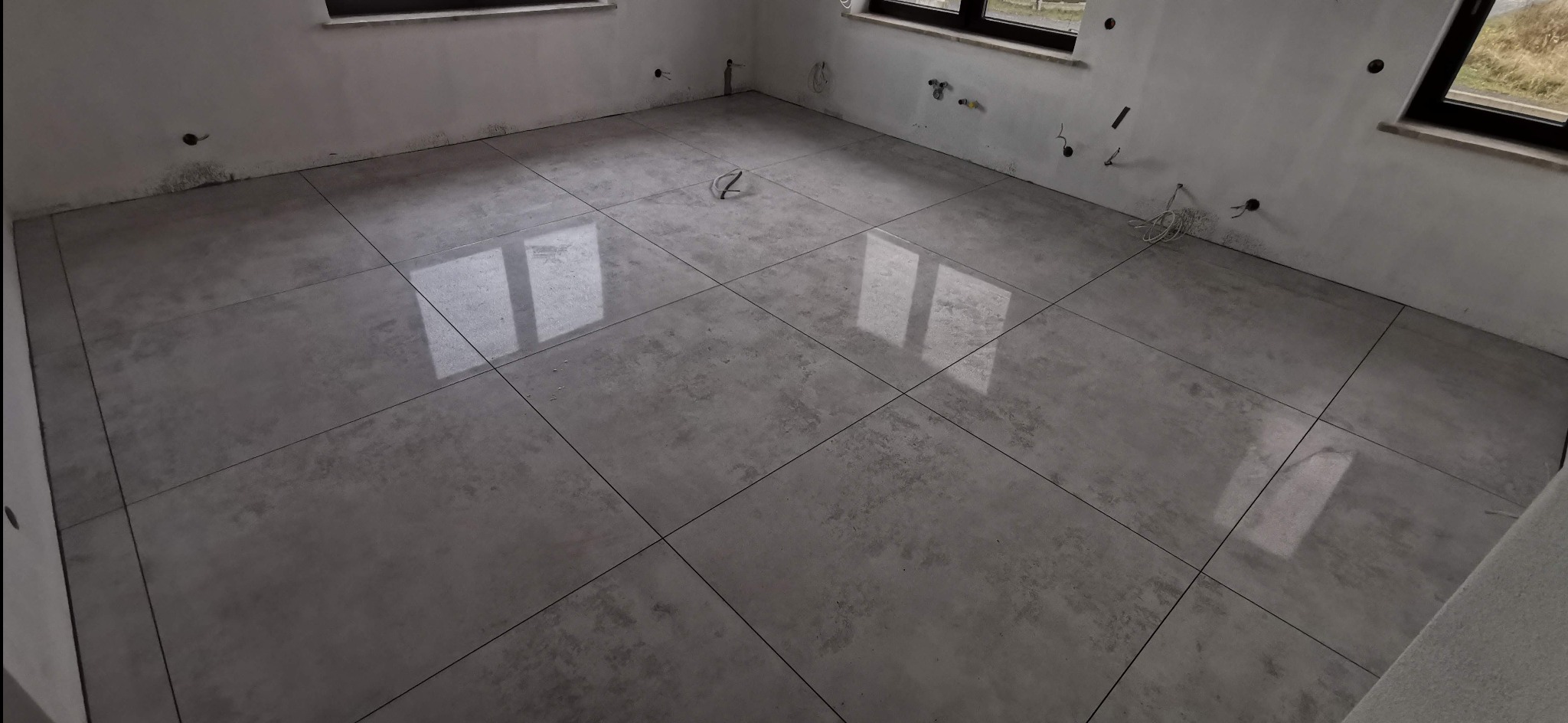 Wykończona podłoga z szarych, matowych płytek w surowym wnętrzu. Widoczne otwory instalacyjne w ścianach i refleksy światła na płytkach. Minimalistyczny design.