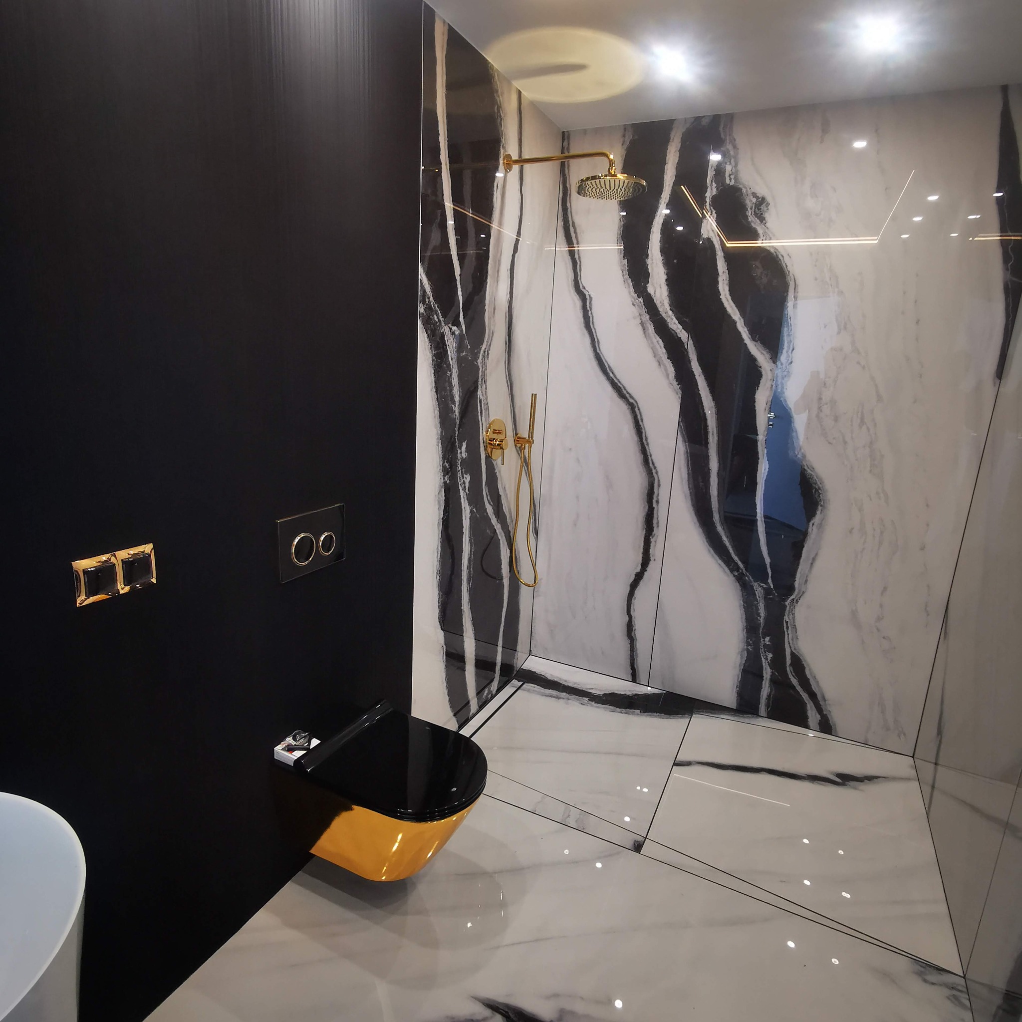 Elegancka łazienka z czarną ścianą, złotą armaturą i toaletą. Ściany i podłoga wyłożone płytkami imitującymi marmur z wyraźnym wzorem.