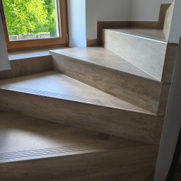 BestBud - Schody wykończone drewnopodobnymi panelami, widoczne stopnie z profilami antypoślizgowymi i fragment okna z widokiem na zieleń. Estetyczne i funkcjonalne rozwiązanie.