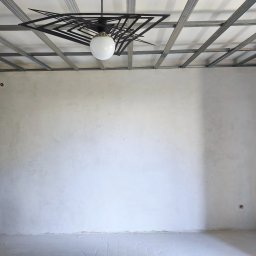 BestBud - Wnętrze w trakcie prac wykończeniowych: szpachlowane ściany, metalowa konstrukcja sufitu podwieszanego i nowoczesna lampa wisząca. Widoczne puszki elektryczne.
