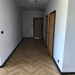 BestBud - Korytarz po remoncie: parkiet w jodełkę, białe ściany z czarnymi listwami przypodłogowymi i ościeżnicami, nowoczesna lampa wisząca, dwoje drzwi.