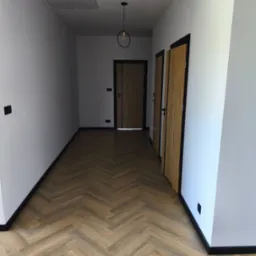 Korytarz po remoncie: parkiet w jodełkę, białe ściany z czarnymi listwami przypodłogowymi i ościeżnicami, nowoczesna lampa wisząca, dwoje drzwi.