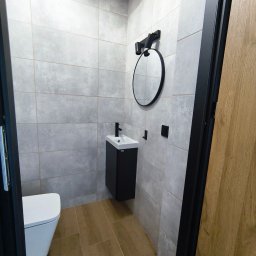 BestBud - Nowoczesna łazienka w szarościach z drewnopodobną podłogą. Umywalka z czarną szafką, okrągłe lustro i wisząca toaleta. Minimalistyczny design.
