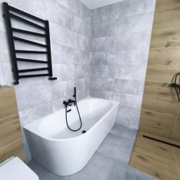 BestBud - Nowoczesna łazienka z wolnostojącą wanną, grafitową armaturą i grzejnikiem drabinkowym na tle szarych płytek. Uwagę zwraca drewnopodobna okładzina.