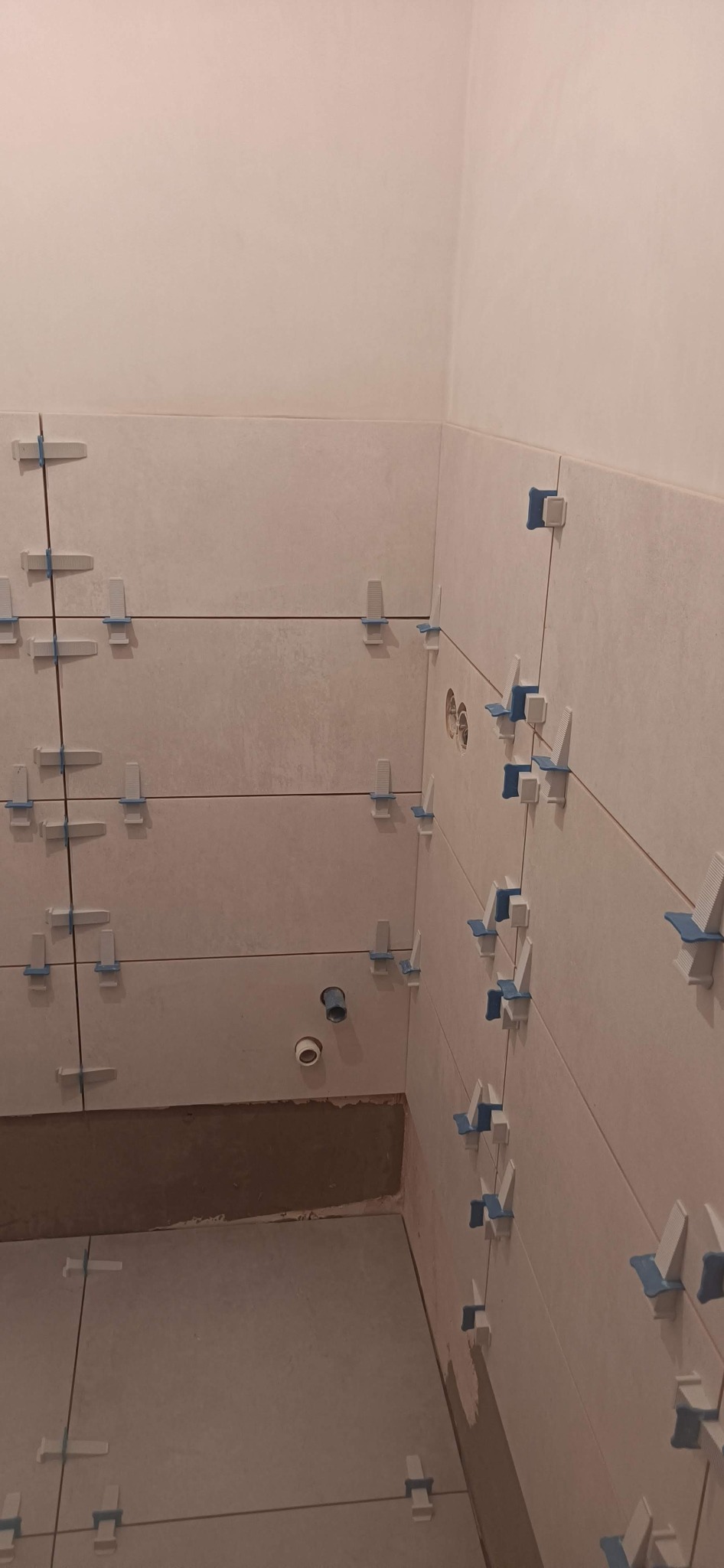 Ściana i podłoga w trakcie układania płytek ceramicznych z użyciem klipsów poziomujących, widoczne otwory na instalacje. Wnętrze łazienki w trakcie wykańczania.