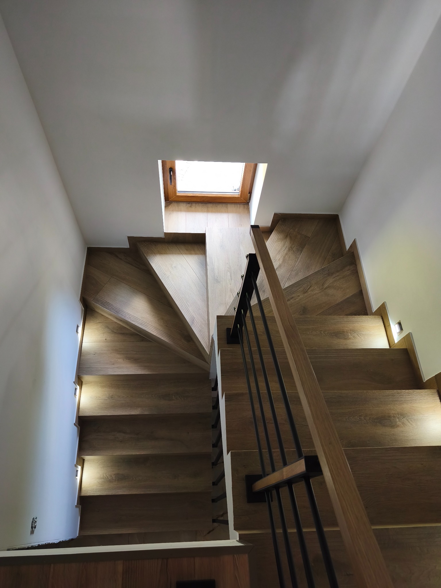 Nowoczesne schody drewniane z oświetleniem LED wzdłuż ściany, widok z góry na kręte stopnie i okno dachowe, minimalistyczne balustrady.