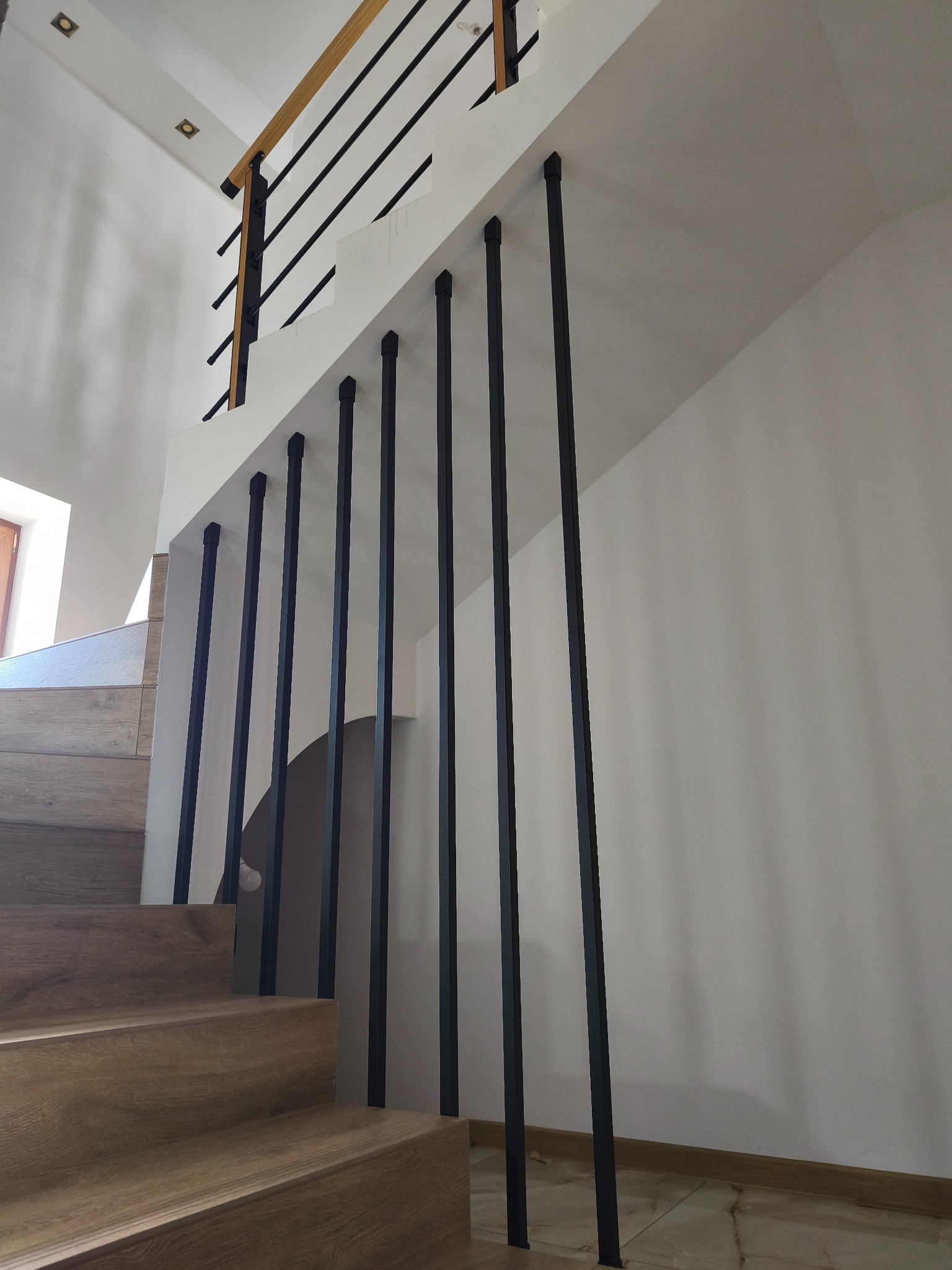 Nowoczesna balustrada schodowa z czarnymi, pionowymi słupkami i drewnianym poręczą, widok z dołu, jasne wnętrze.