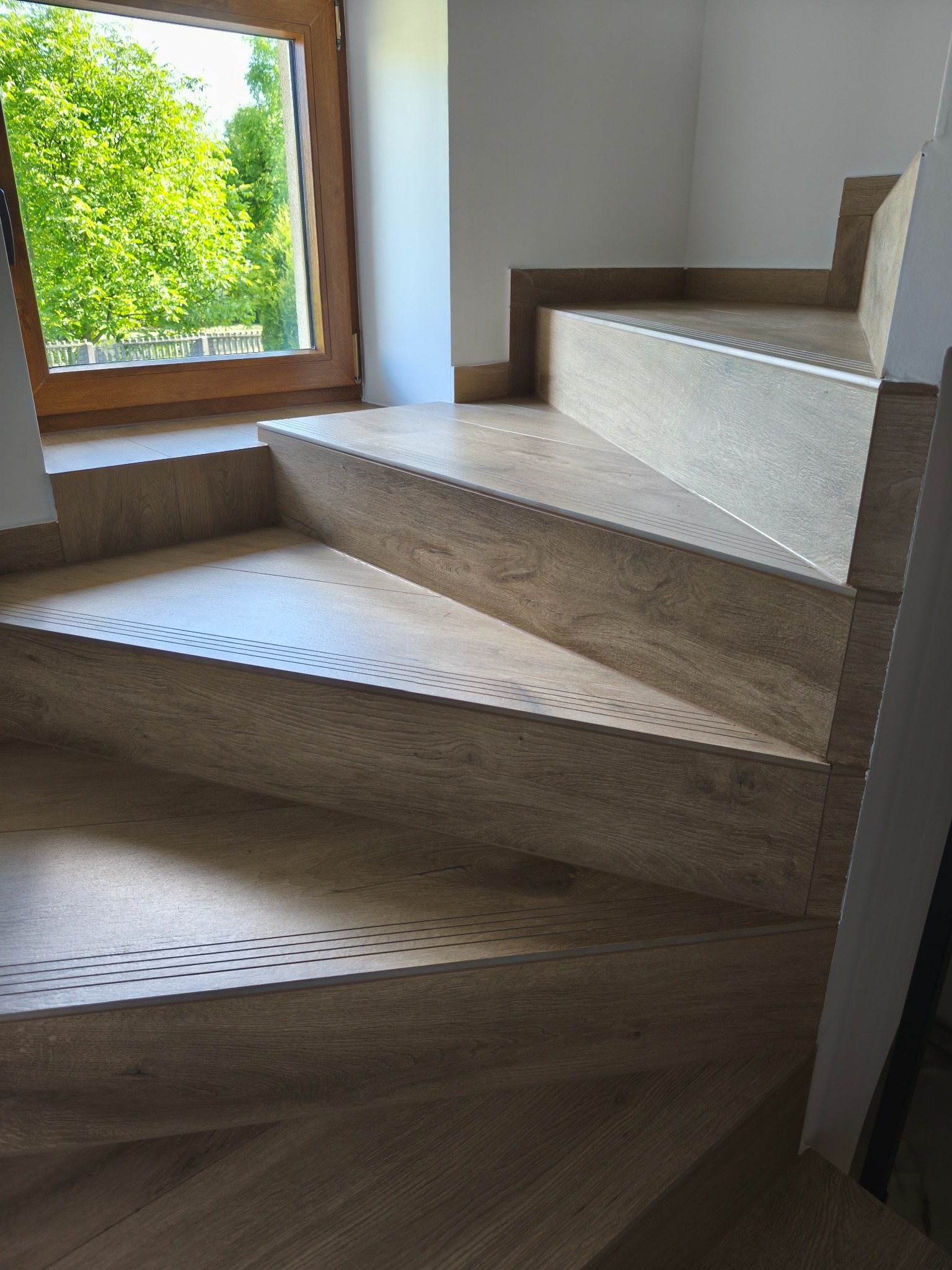 Schody wykończone drewnopodobnymi panelami, widoczne stopnie z profilami antypoślizgowymi i fragment okna z widokiem na zieleń. Estetyczne i funkcjonalne rozwiązanie.