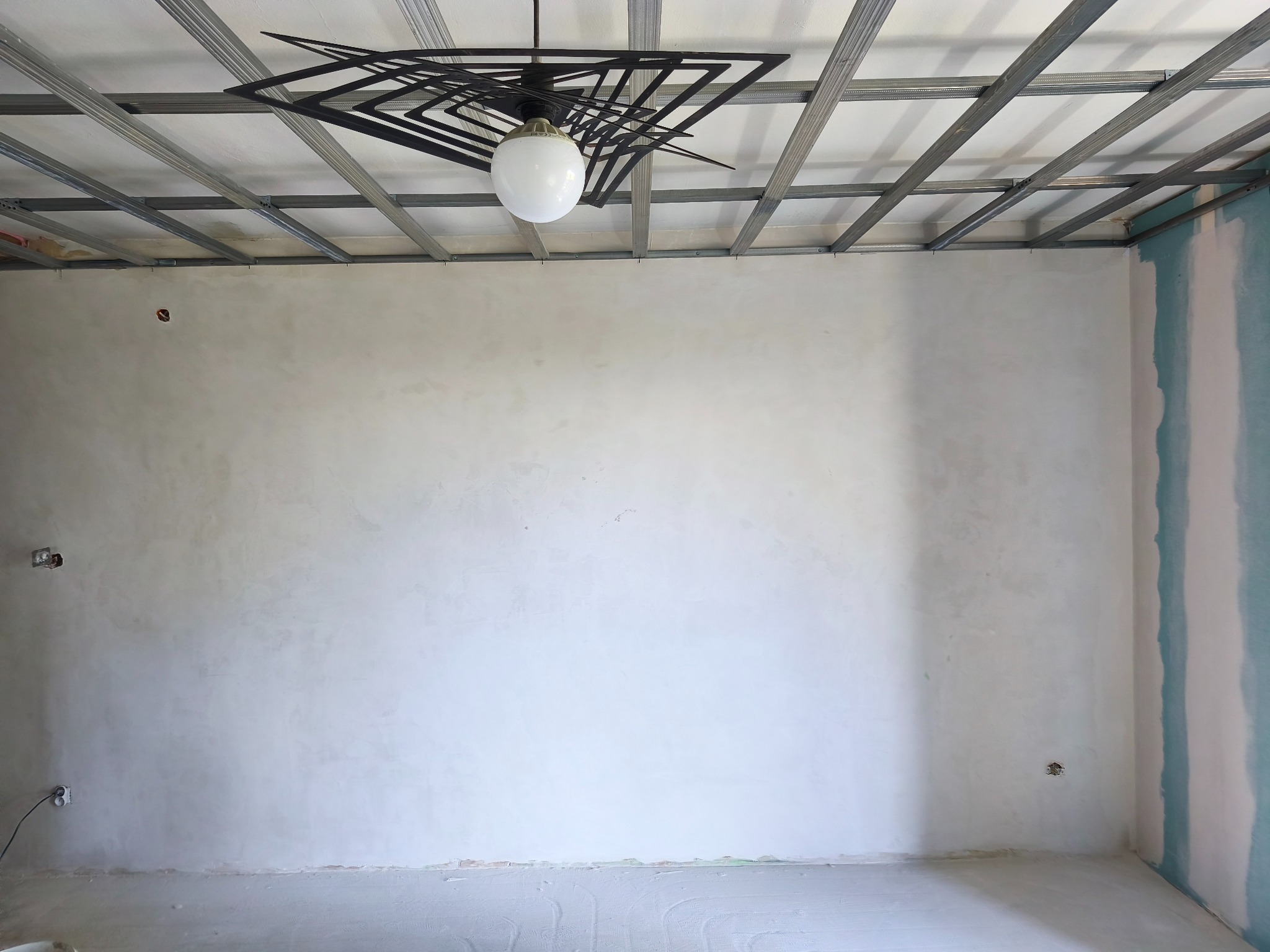 Wnętrze w trakcie prac wykończeniowych: szpachlowane ściany, metalowa konstrukcja sufitu podwieszanego i nowoczesna lampa wisząca. Widoczne puszki elektryczne.